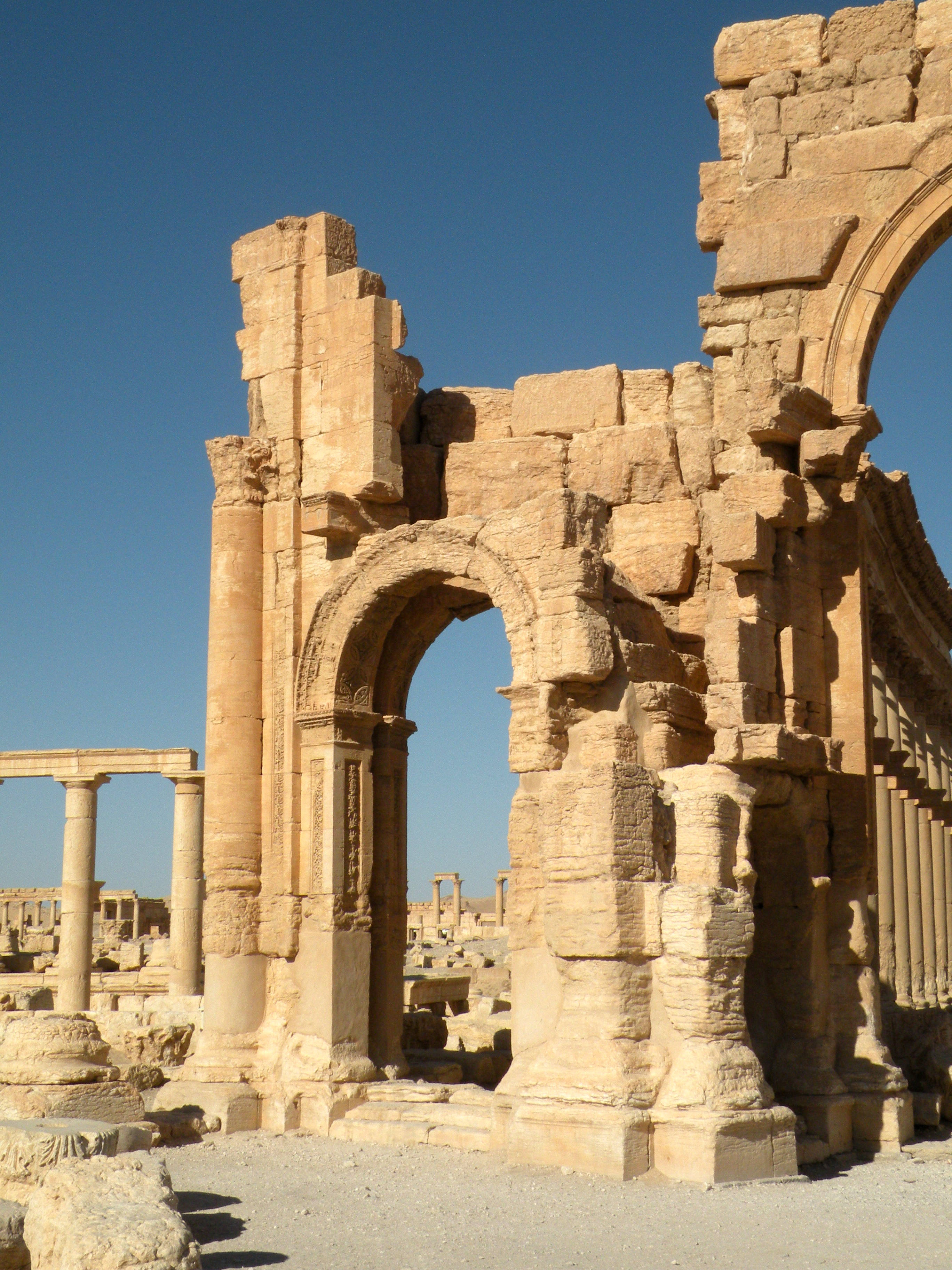 Syria 桌布 壁紙 — 陽光照射下的古老石頭廢墟，有拱形門廊和斷裂的柱子，在沙漠景觀中映襯著清澈的藍天。