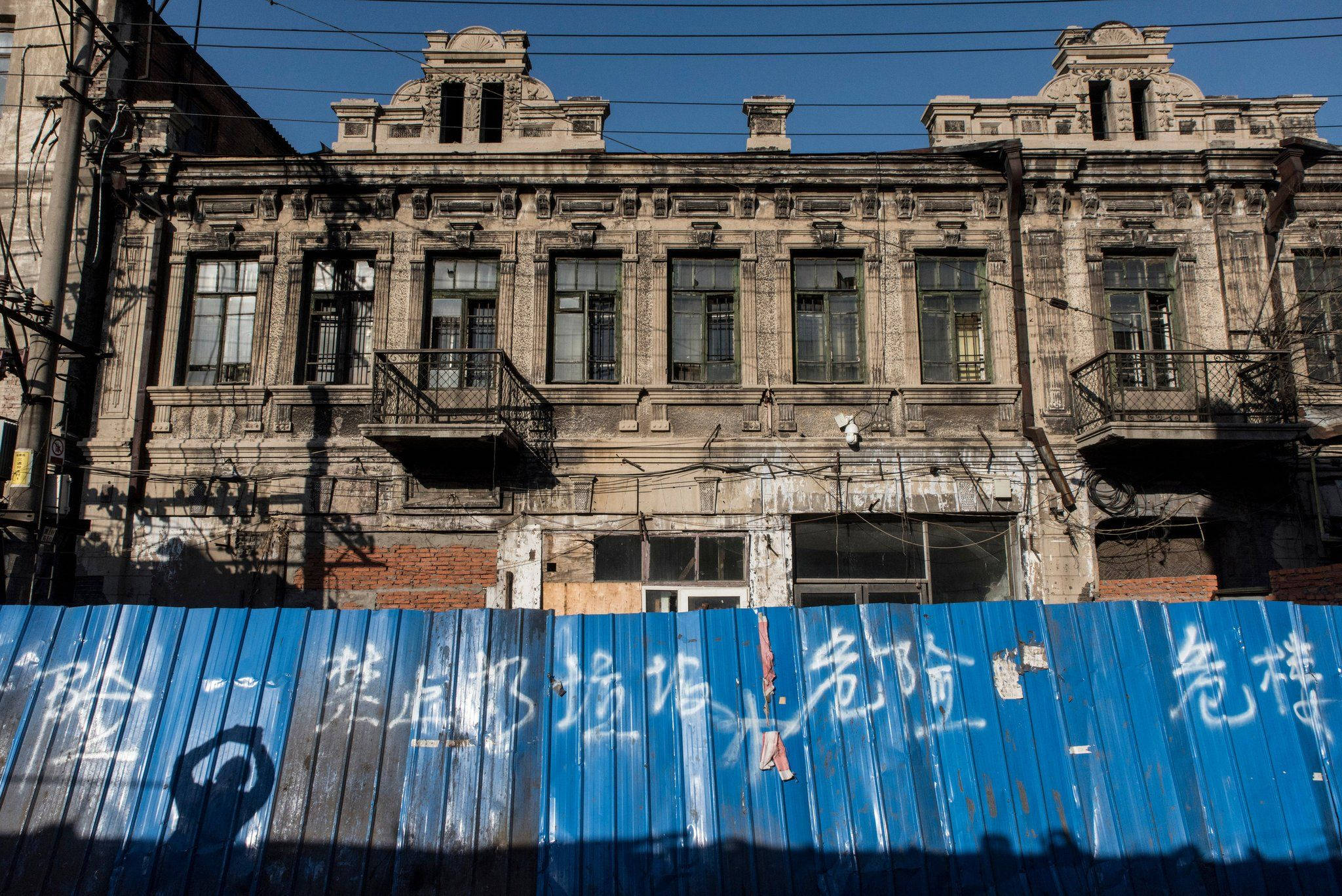 Harbin 桌布 壁紙 — 陽光照射下破舊的華麗建築，前景有藍色波紋柵欄投下長長的影子。