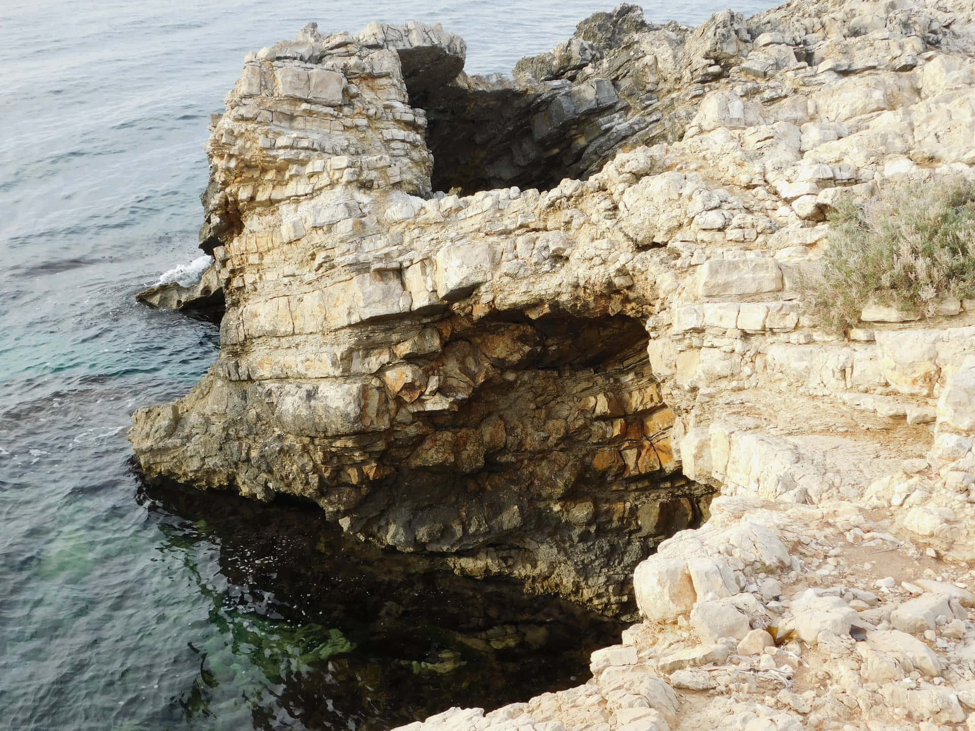 Sea with rocks 壁紙 — 陽光照射的層狀岩層，帶有天然拱門，伸入碧綠色的海洋，伴隨著輕柔的波浪。