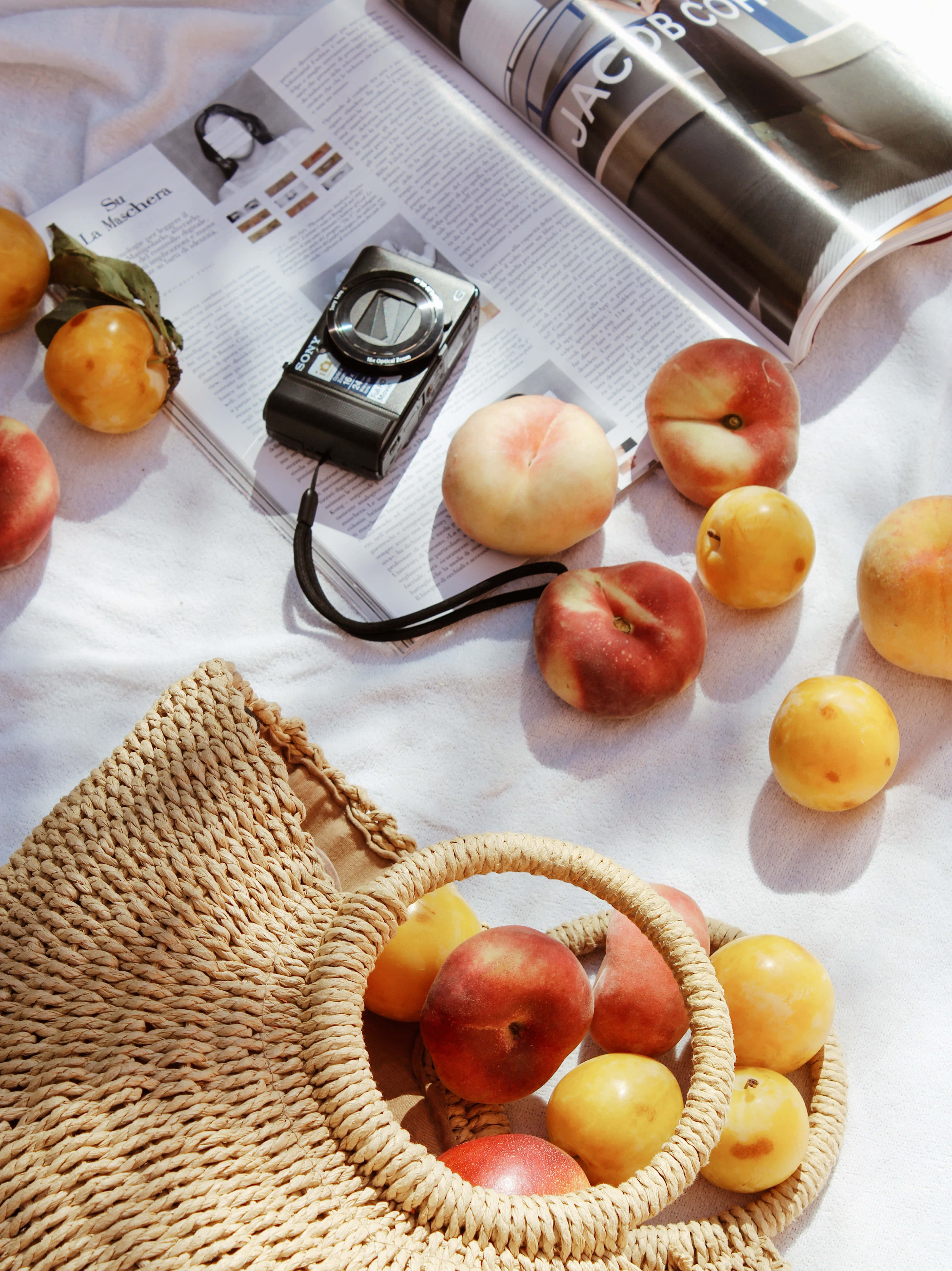 Peaches 桌布 壁紙 — 陽光照射的桃子和李子從編織籃中溢出，旁邊是打開的雜誌和復古相機，放在白色布料上。