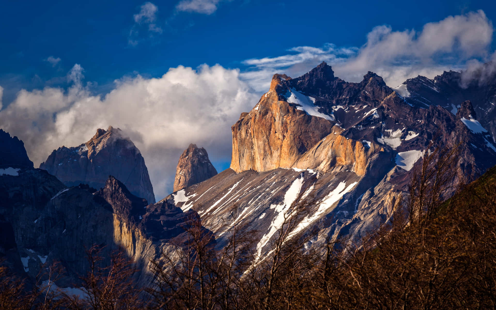 Patagonia 桌布 壁紙 — 陽光照射的花崗岩山峰穿透戲劇性的雲層，覆蓋著雪的山脊，前景是光禿禿的樹木。