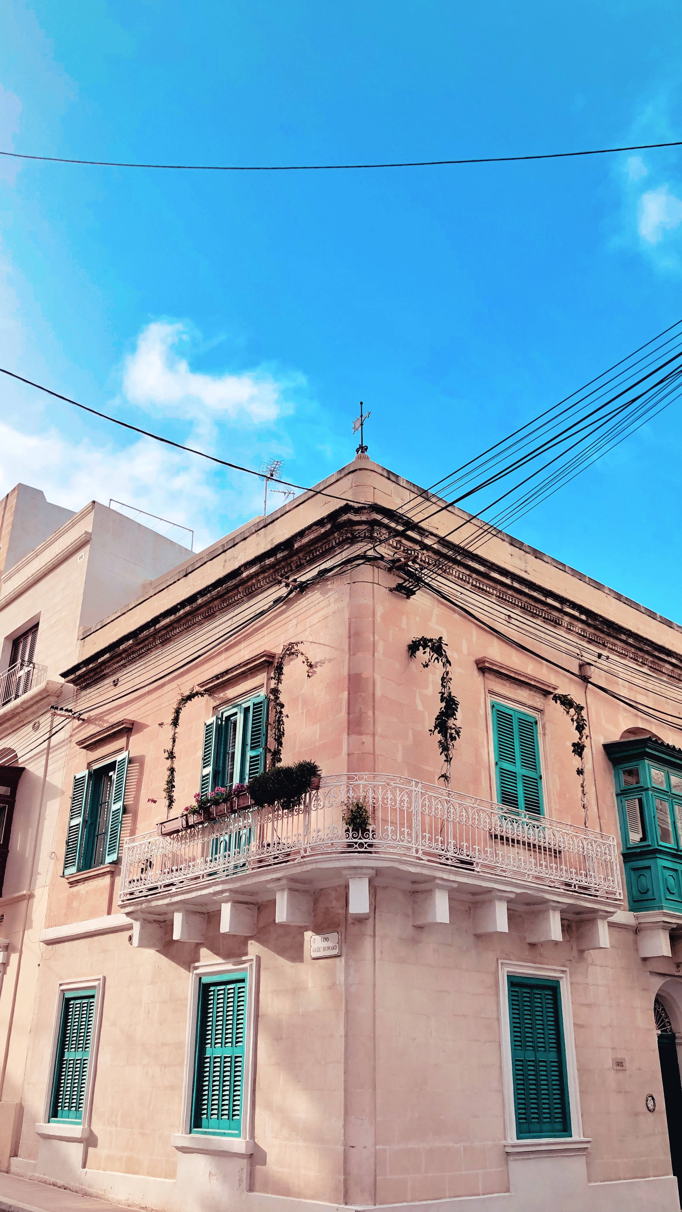 Malta 桌布 壁紙 — 陽光照射的角落建築，桃色牆壁搭配青綠色百葉窗，在明亮藍天與薄雲下。