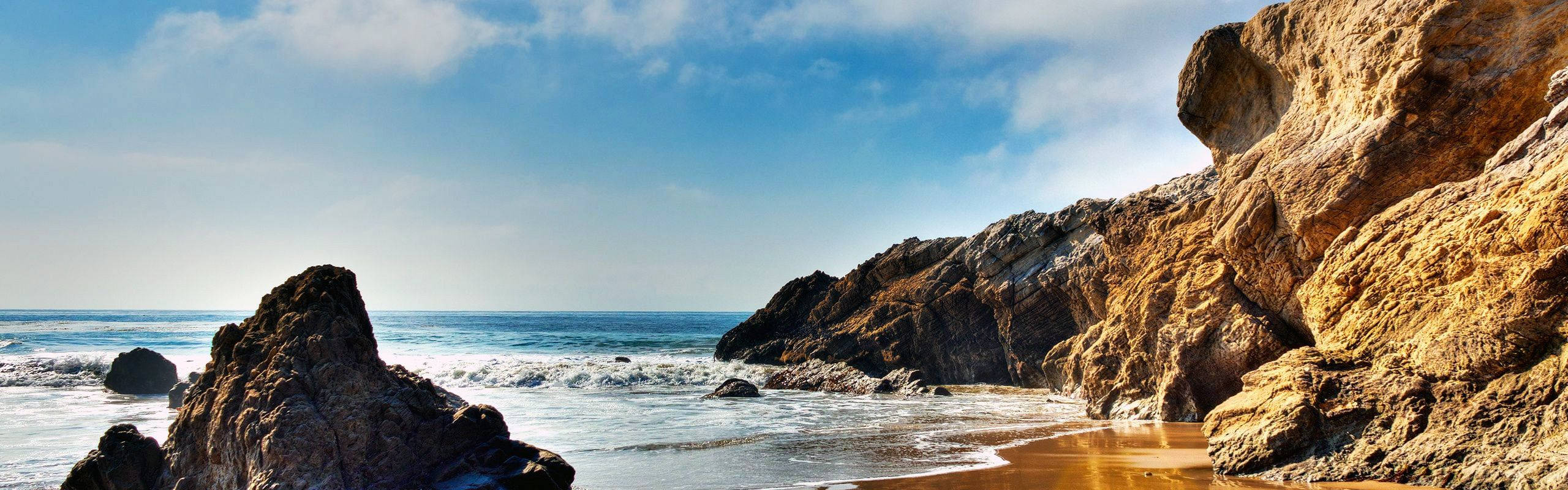 Malibu 桌布 壁紙 — 陽光照射的金色懸崖環繞著沙灘，海浪在清澈的藍天下拍打著堅固的岩石。