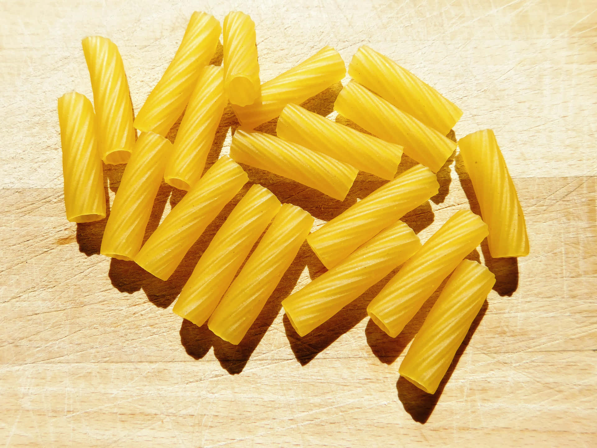 Pasta 壁紙 — 陽光照射的黃色螺紋粉管散落在淺色木砧板上，投射出清晰的陰影。