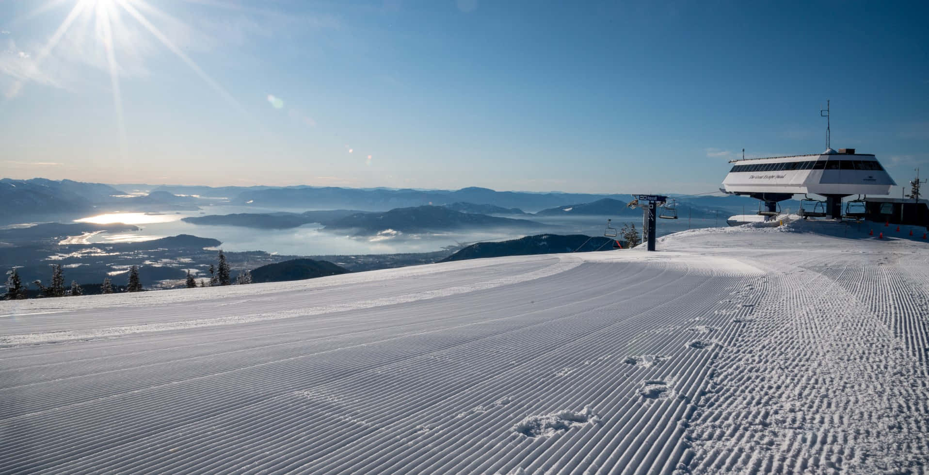 Snow Zoom 桌布 壁紙 — 陽光照耀、白雪覆蓋的滑雪坡道，設有修整過的雪道和纜車站，俯瞰霧氣瀰漫的山谷和遠山。