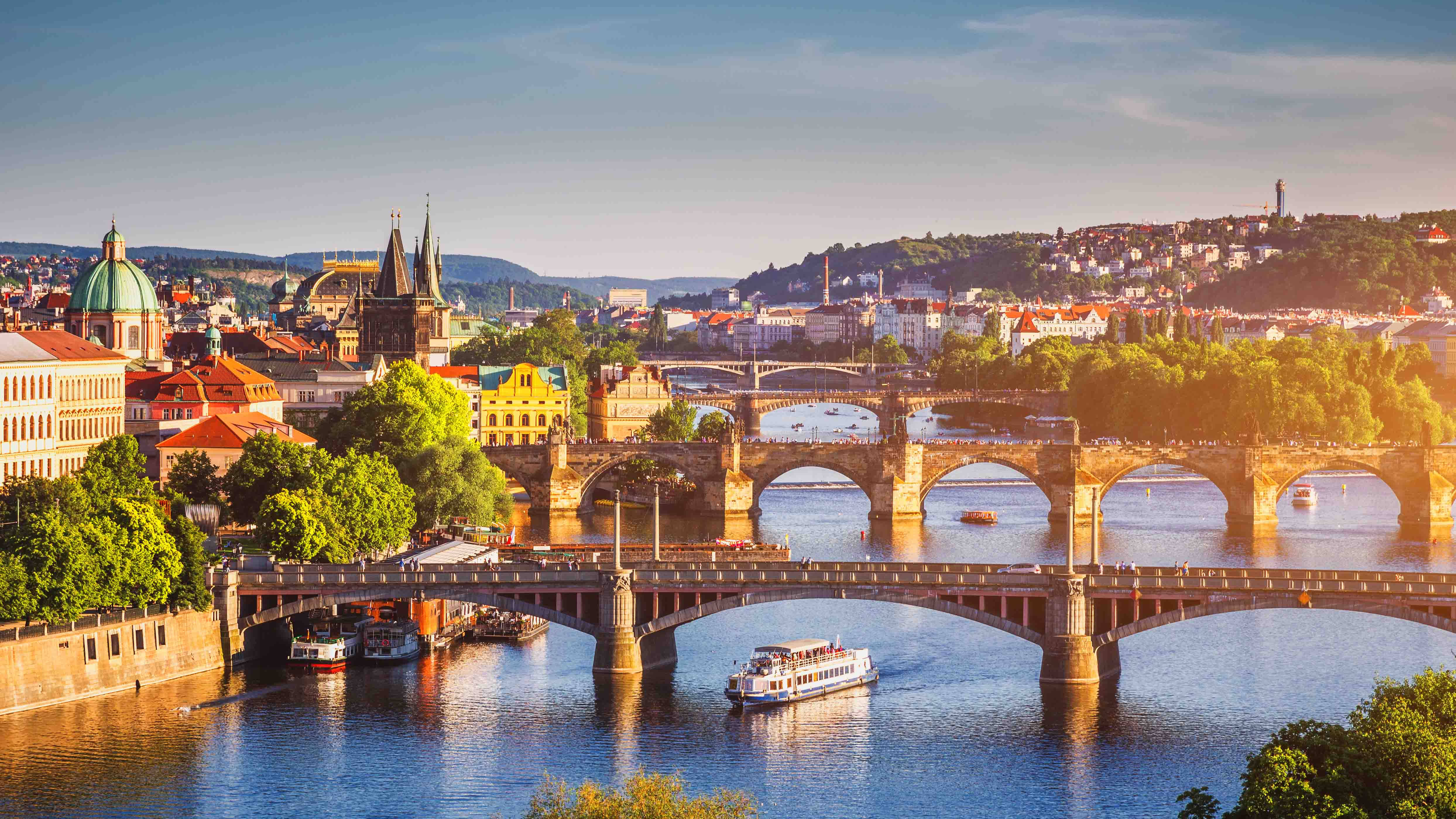 Europe 桌布 壁紙 — 陽光照耀下的布拉格橋樑橫跨河流，河上有船隻，歷史建築和綠色山丘框出充滿活力的城市景觀。
