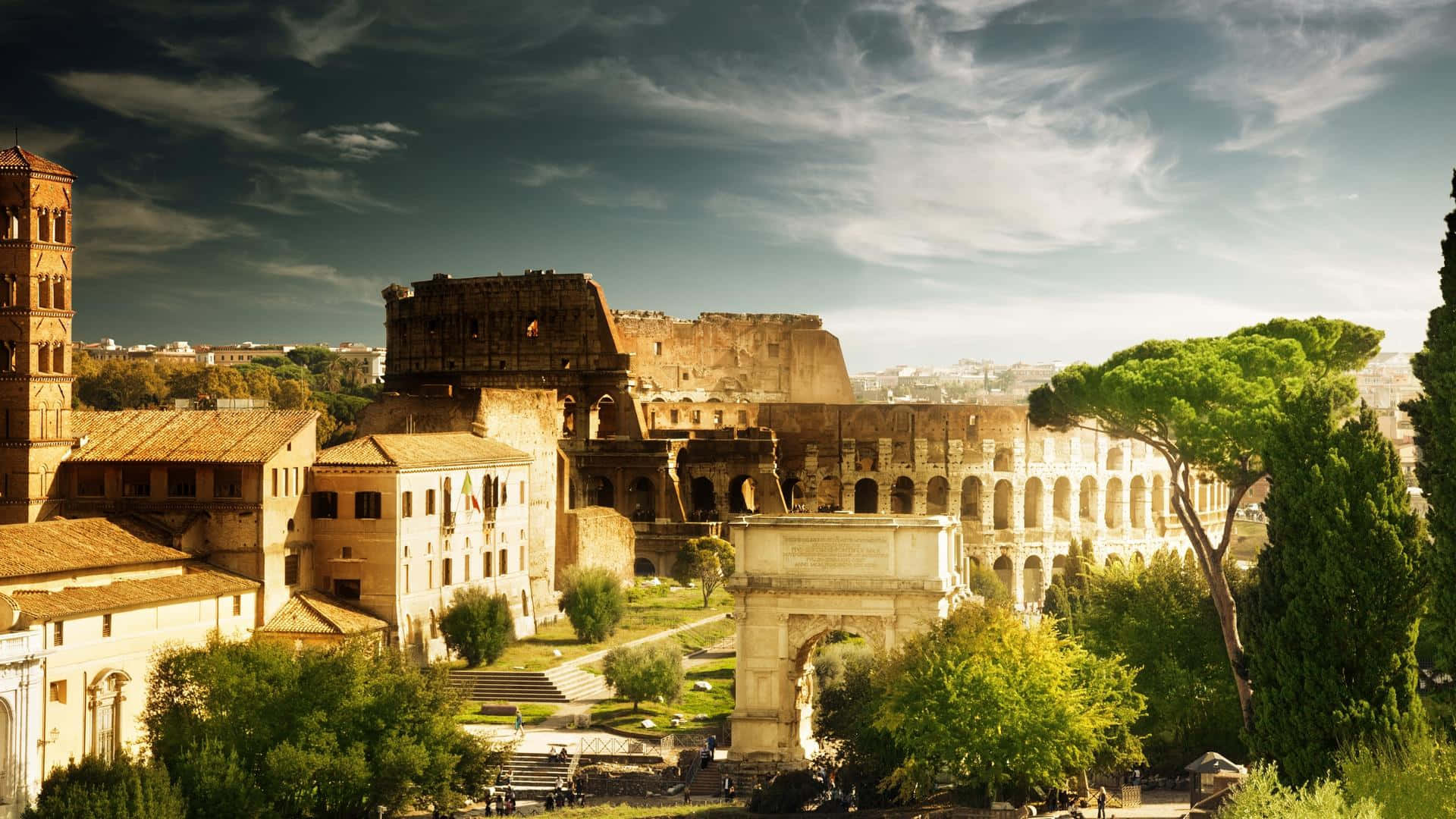 Ancient Roman Desktop 桌布 壁紙 — 陽光照耀下的羅馬競技場及周圍的古代遺跡，被茂密的綠樹環繞，背景是戲劇性的多雲天空。
