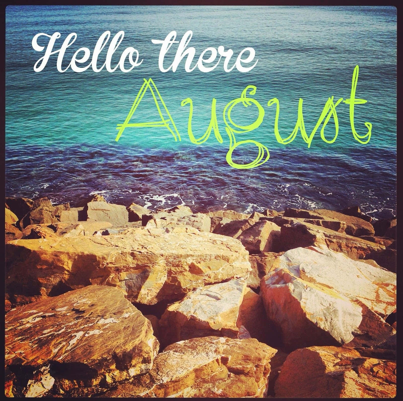 August 桌布 壁紙 — 陽光照耀的岩石海岸在明亮的夏日天空下與碧綠的海浪相遇，帶有「你好八月」文字疊加。