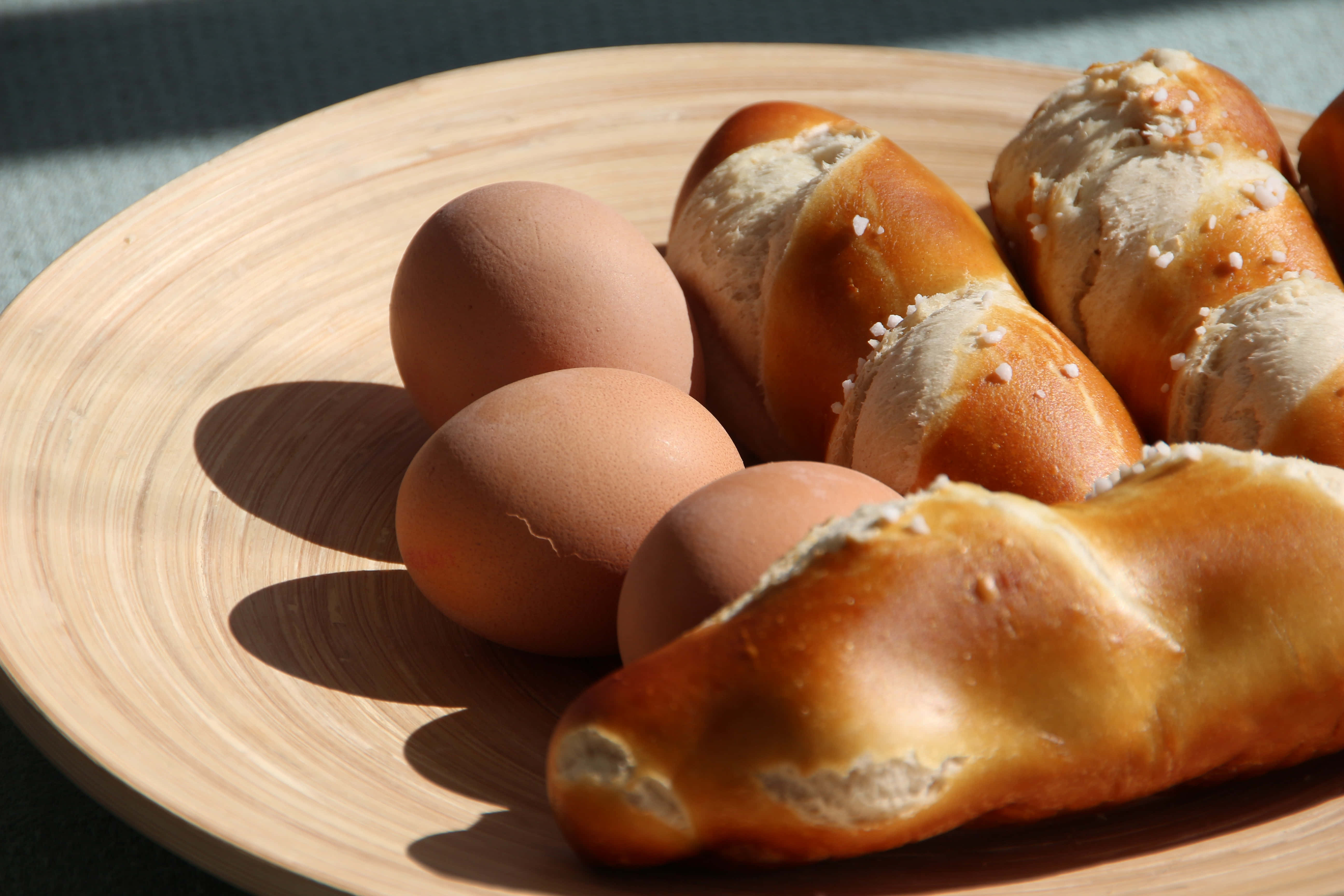 Brown Eggs 桌布 壁紙 — 陽光照耀的棕色雞蛋和金黃色鹽味蝴蝶酥卷放在木盤上，投下清晰的陰影。