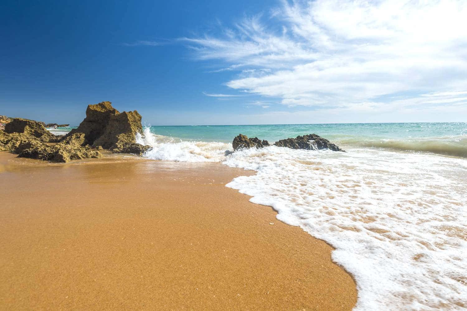 Spanish Beach 桌布 壁紙 — 陽光照耀的海浪拍打著金色沙灘，旁邊是崎嶇的海岸岩石，天空湛藍，雲朵輕柔。