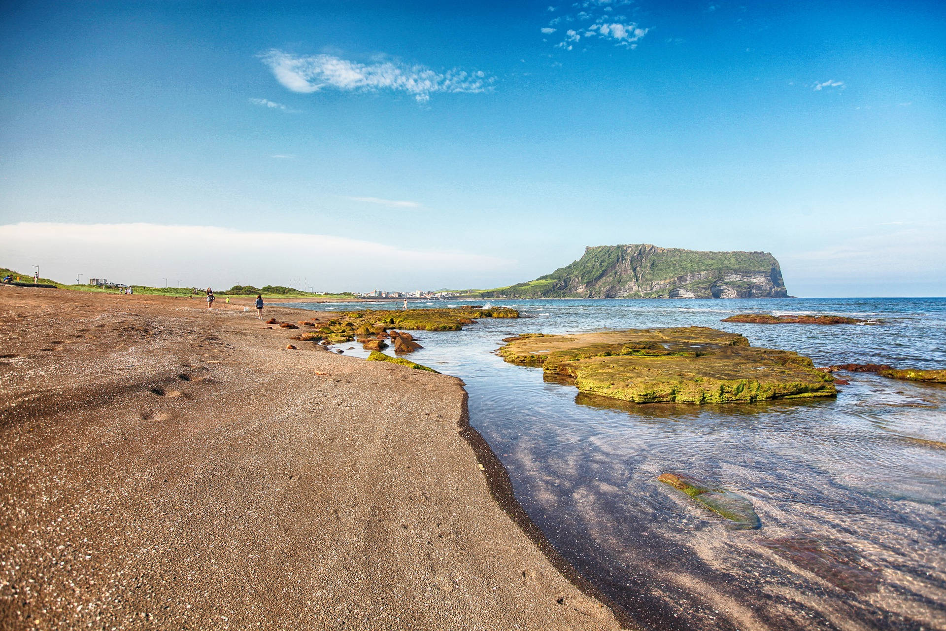 Jeju 桌布 壁紙 — 陽光照耀的海灘，濕潤的岩石和淺水通向遠處的綠色懸崖，天空清澈湛藍。