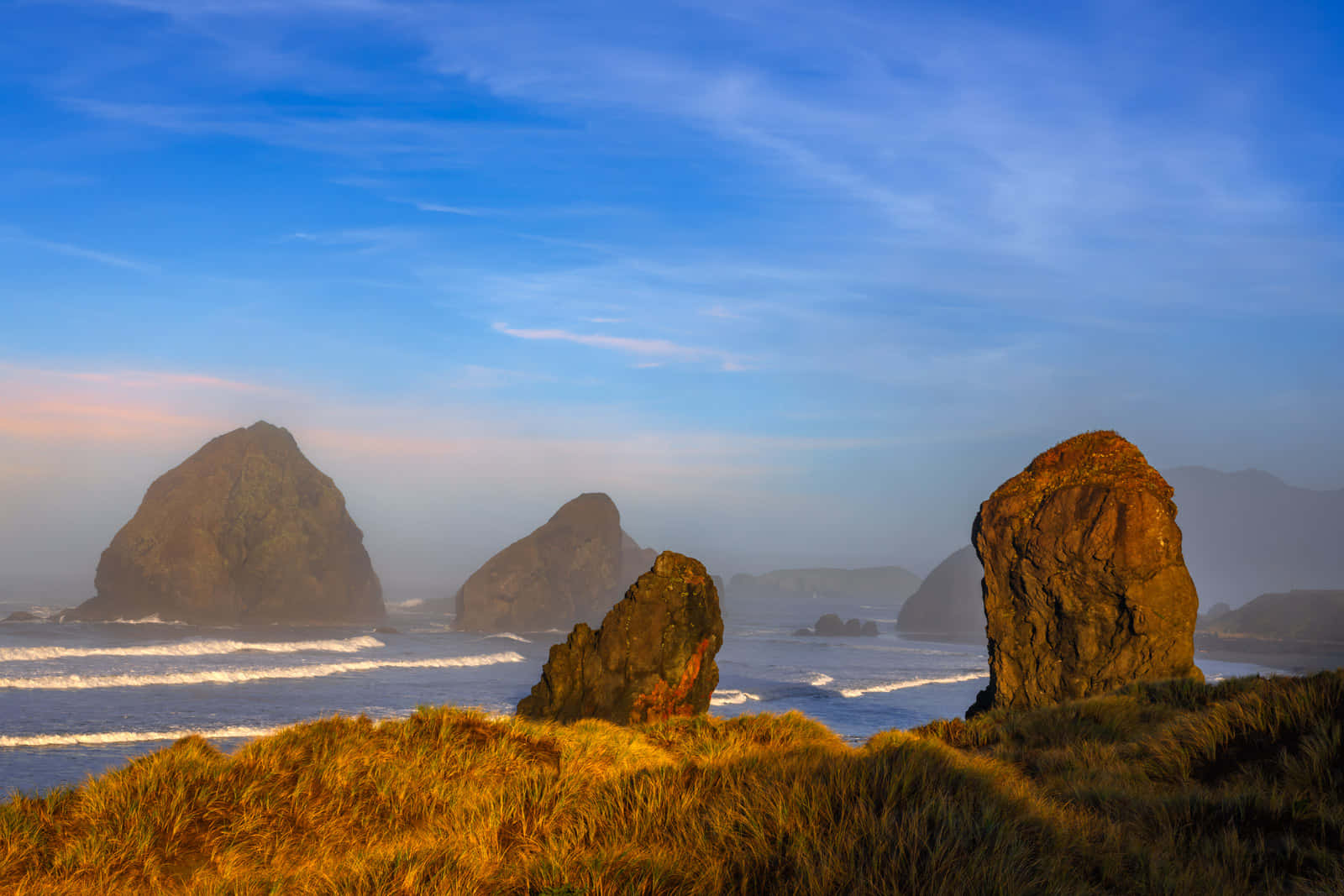 Oregon Coast 桌布 壁紙 — 陽光照耀的草坡懸崖俯瞰著壯觀的海蝕柱和拍岸的浪花，天空柔和藍色，飄著縷縷白雲。
