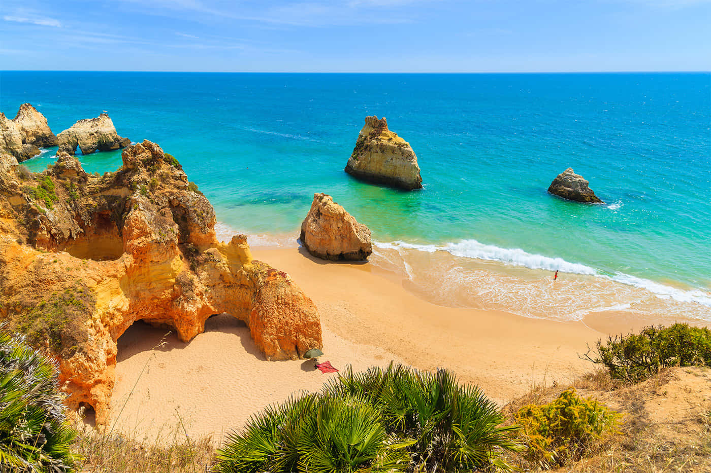 Portuguese Beach 桌布 壁紙 — 陽光照耀的金色懸崖框住一片碧綠色的海洋，散落著岩石 formations 和一個孤獨的游泳者靠近沙灘。