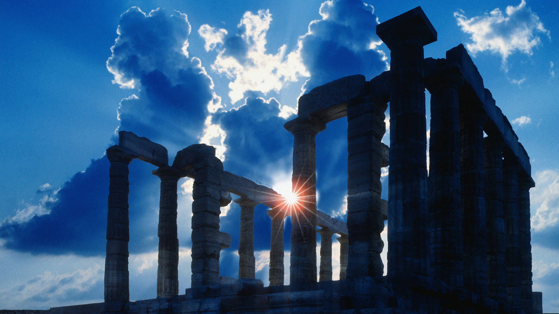 Athens 桌布 壁紙 — 陽光穿透古老石柱，映襯著戲劇性的藍天和蓬鬆的白雲。