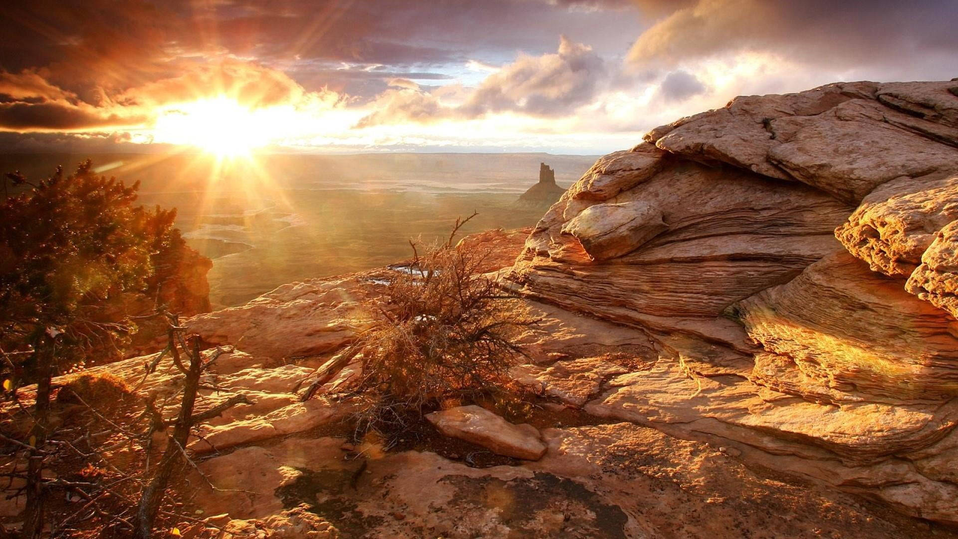 Canyonlands National Park 桌布 壁紙 — 陽光穿透崎嶇峽谷懸崖上空的雲層，將金色光芒灑在層狀岩層和稀疏的沙漠灌木上。