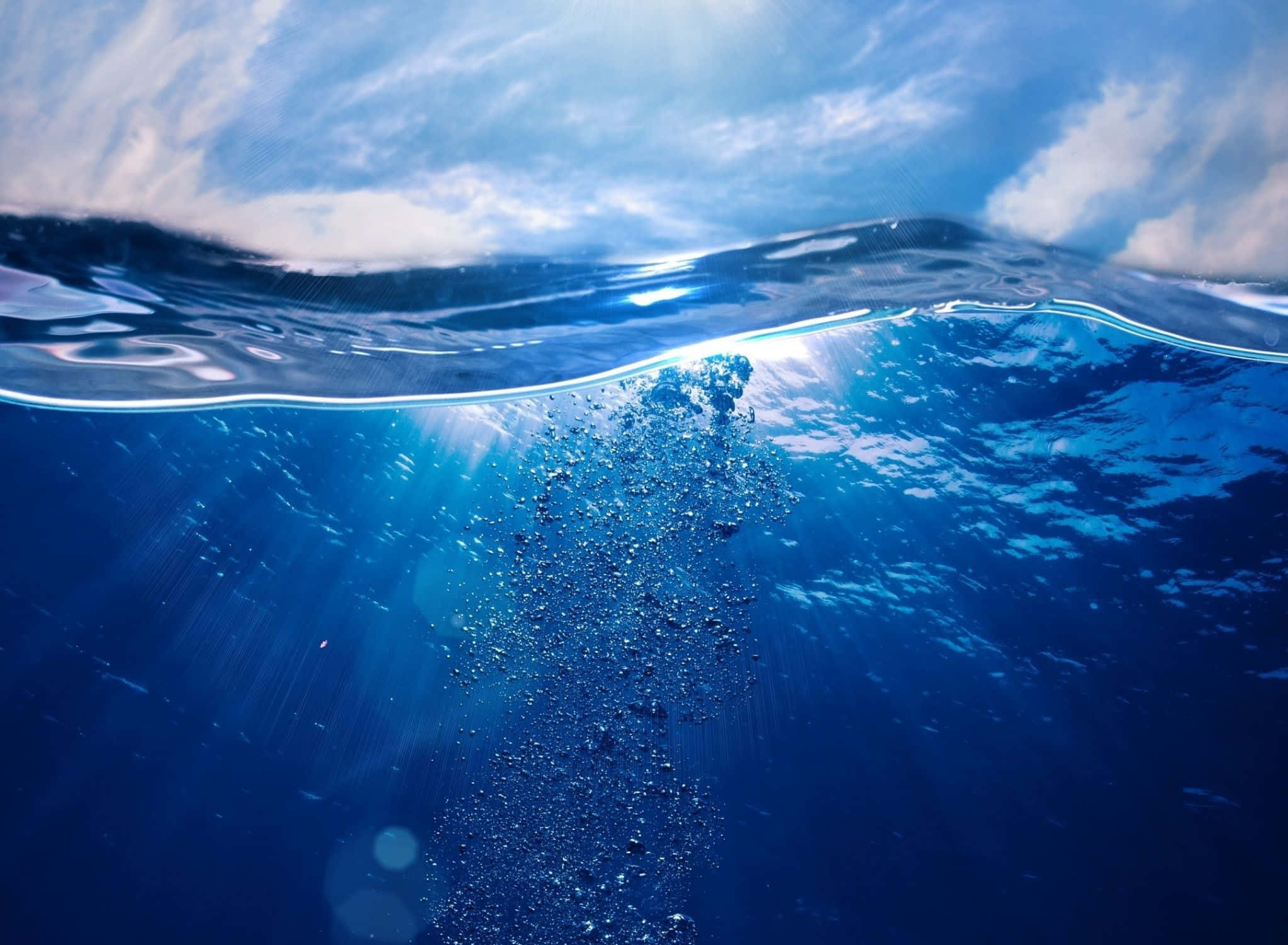 海洋藍 桌布 壁紙 — 陽光穿透海浪，形成一個閃閃發光的水下場景，有上升的氣泡和上方清澈的藍天。
