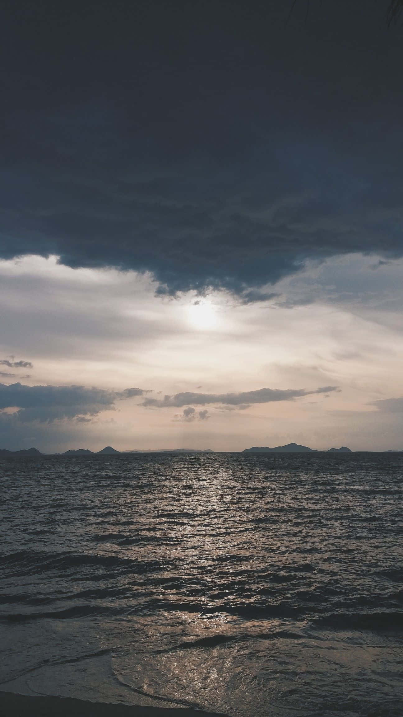 Ocean Aesthetic Phone 桌布 壁紙 — 陽光穿透黑暗的暴風雲，在遠處島嶼附近波光粼粼的海面上投下金色光芒。