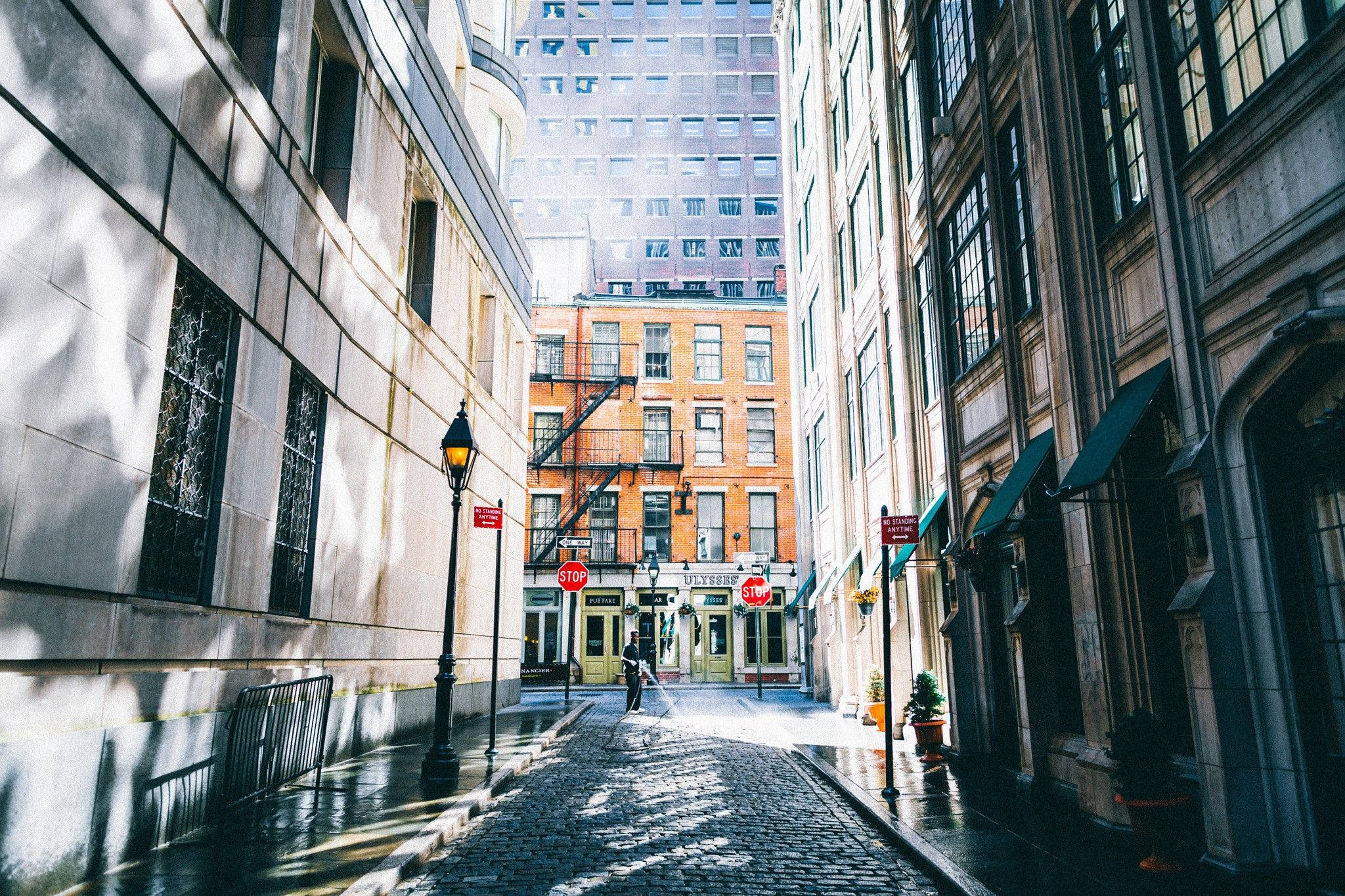 New York Street 桌布 壁紙 — 陽光穿過一條狹窄的鵝卵石小巷，兩旁是歷史建築和遠處的現代摩天大樓。