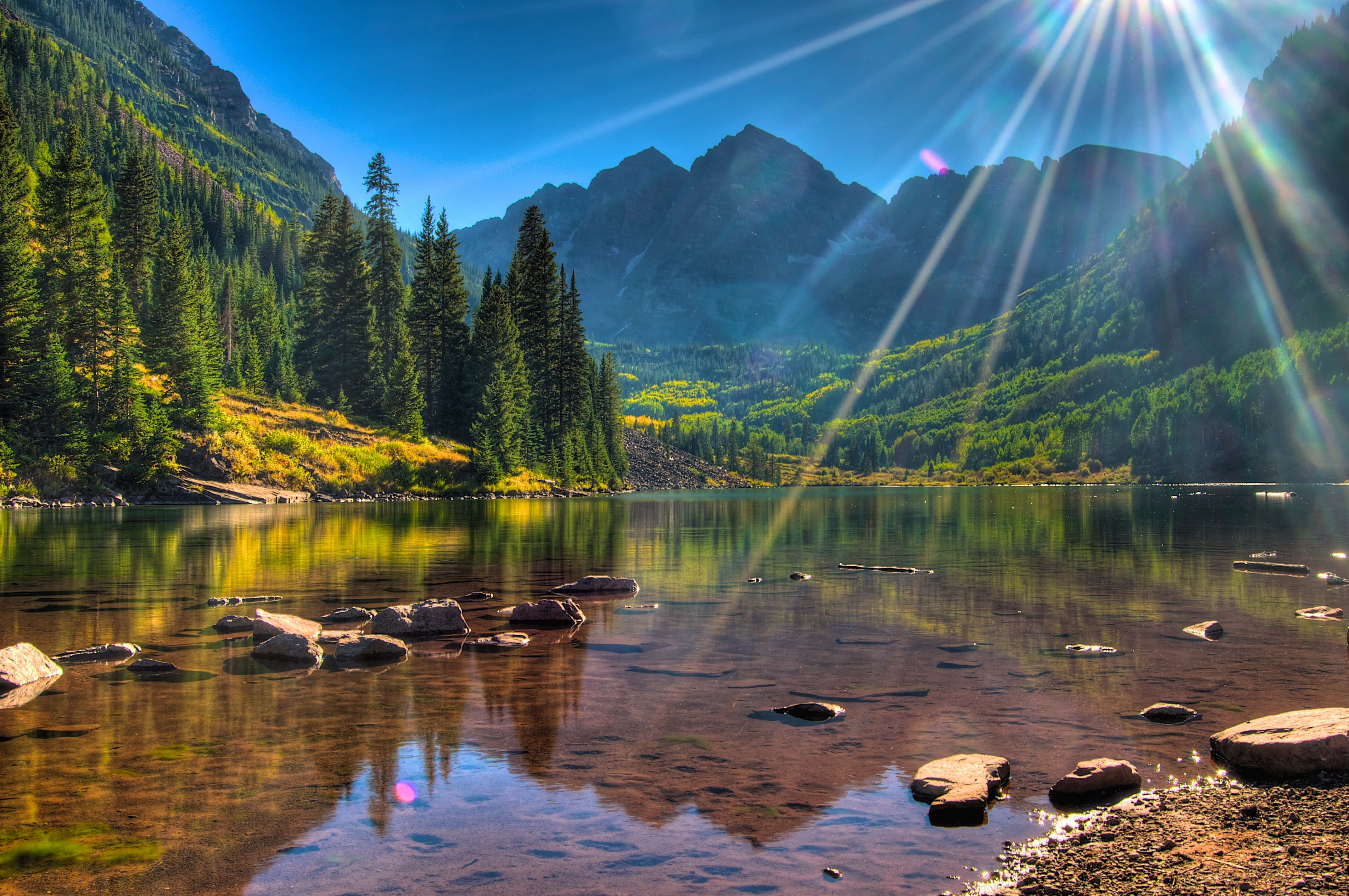 Aspen Colorado 桌布 壁紙 — 陽光穿過山頂，在平靜的湖面上投射出耀眼的光束，映照出常綠森林和岩石海岸。