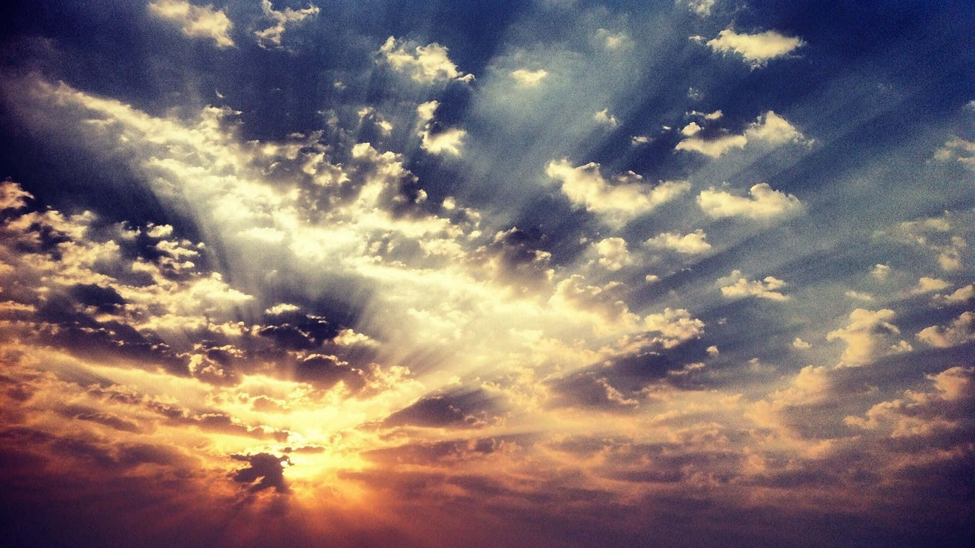 高清 桌布 壁紙 — 陽光穿過戲劇性的雲層，在充滿活力的日落天空中呈現金色光芒和深藍色調。