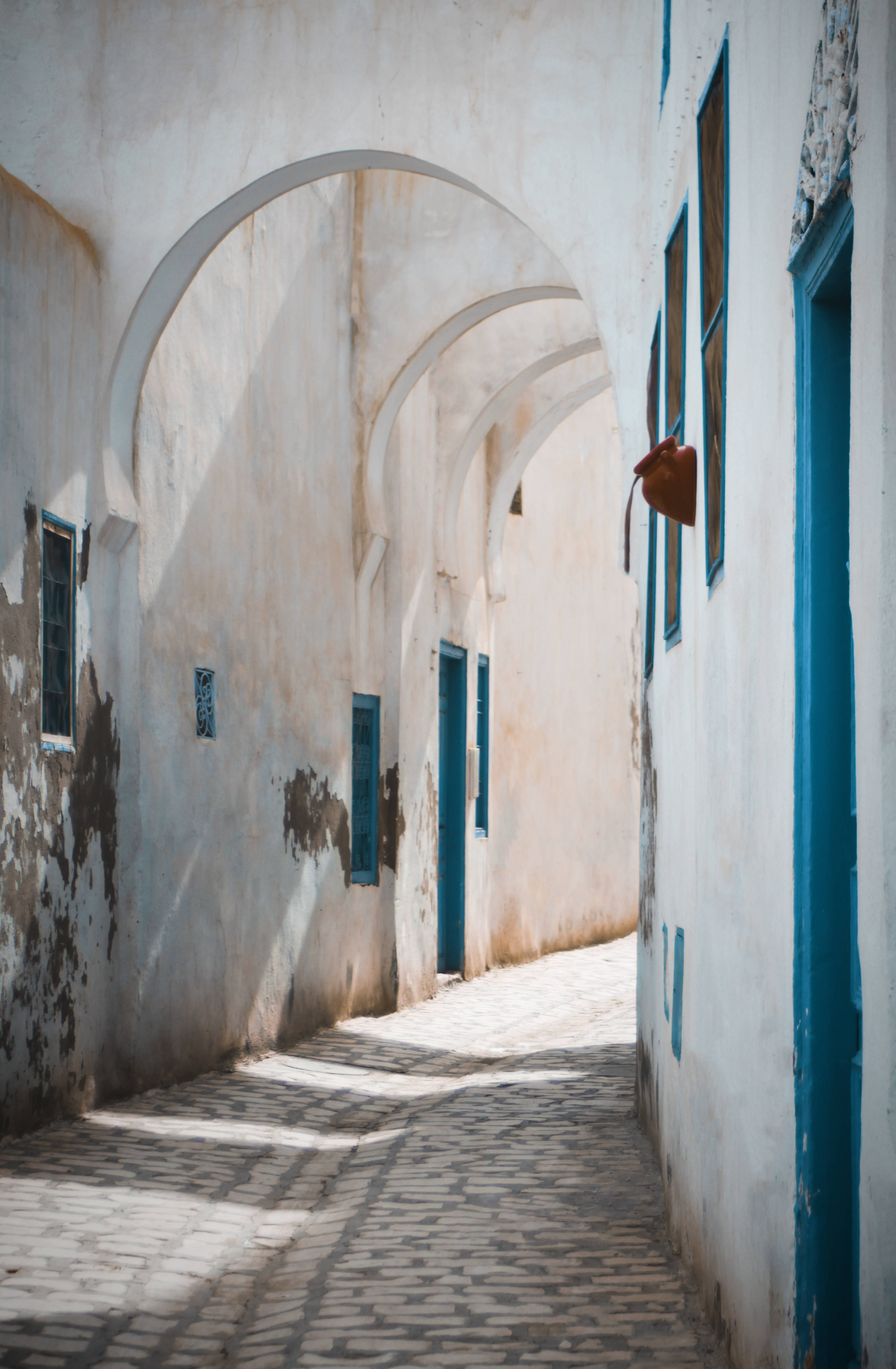 Tunisia 桌布 壁紙 — 陽光穿過拱形門口，在鵝卵石小巷上投下長長的影子，小巷兩旁是風化的白色牆壁和鮮豔的藍色點綴。