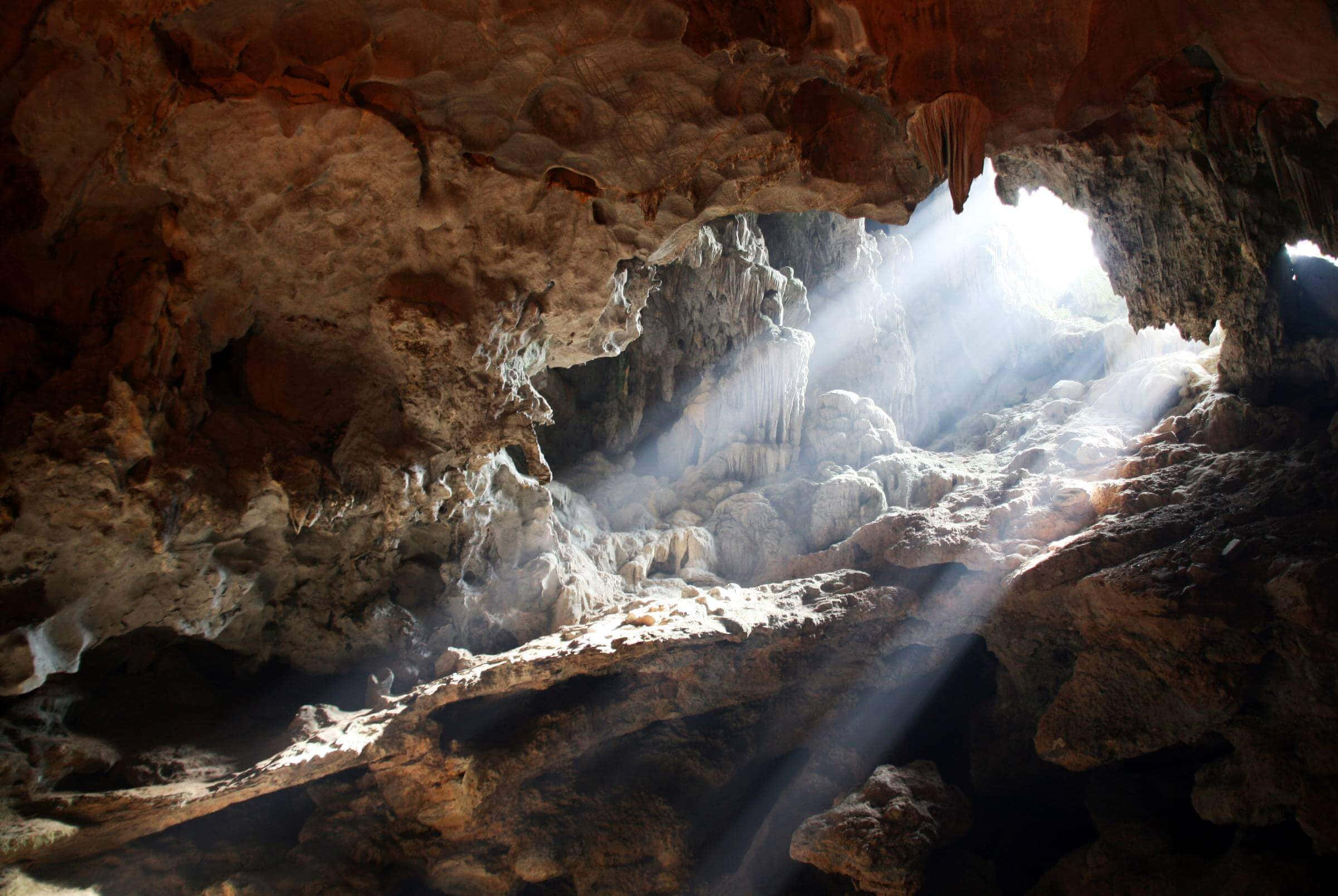 Cave 桌布 壁紙 — 陽光穿過洞穴開口，照亮崎嶇的岩石結構和懸掛的鐘乳石，呈現溫暖的大地色調。