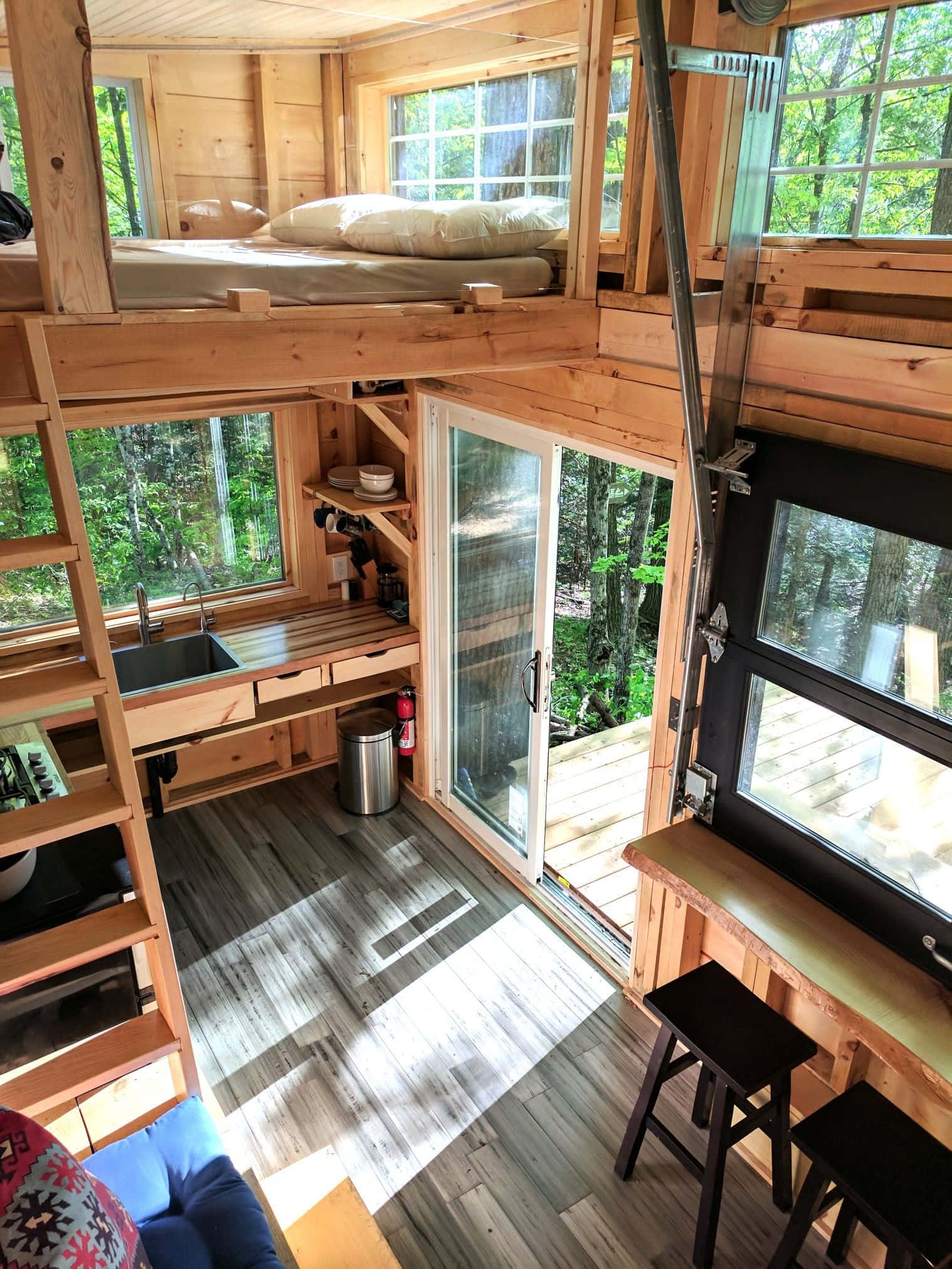 cabin 桌布 壁紙 — 陽光穿過玻璃門，灑進一間舒適的木屋，內有閣樓床、廚房和森林景觀。