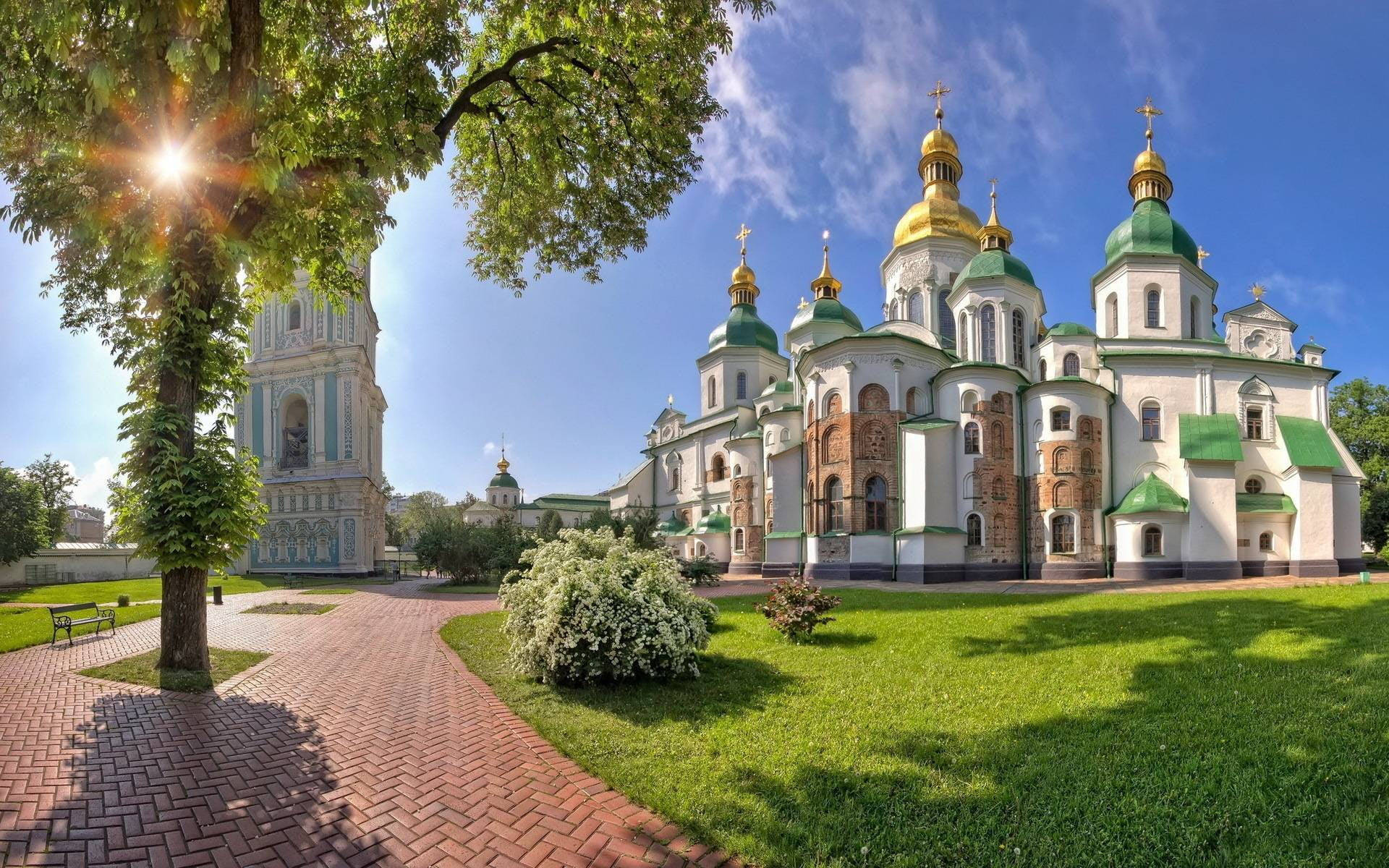 Ukraine 桌布 壁紙 — 陽光穿過茂密的綠樹，在磚砌小徑上投下陰影，小徑通向一座宏偉的大教堂，擁有金色圓頂和白色石牆，在清澈的藍天下。