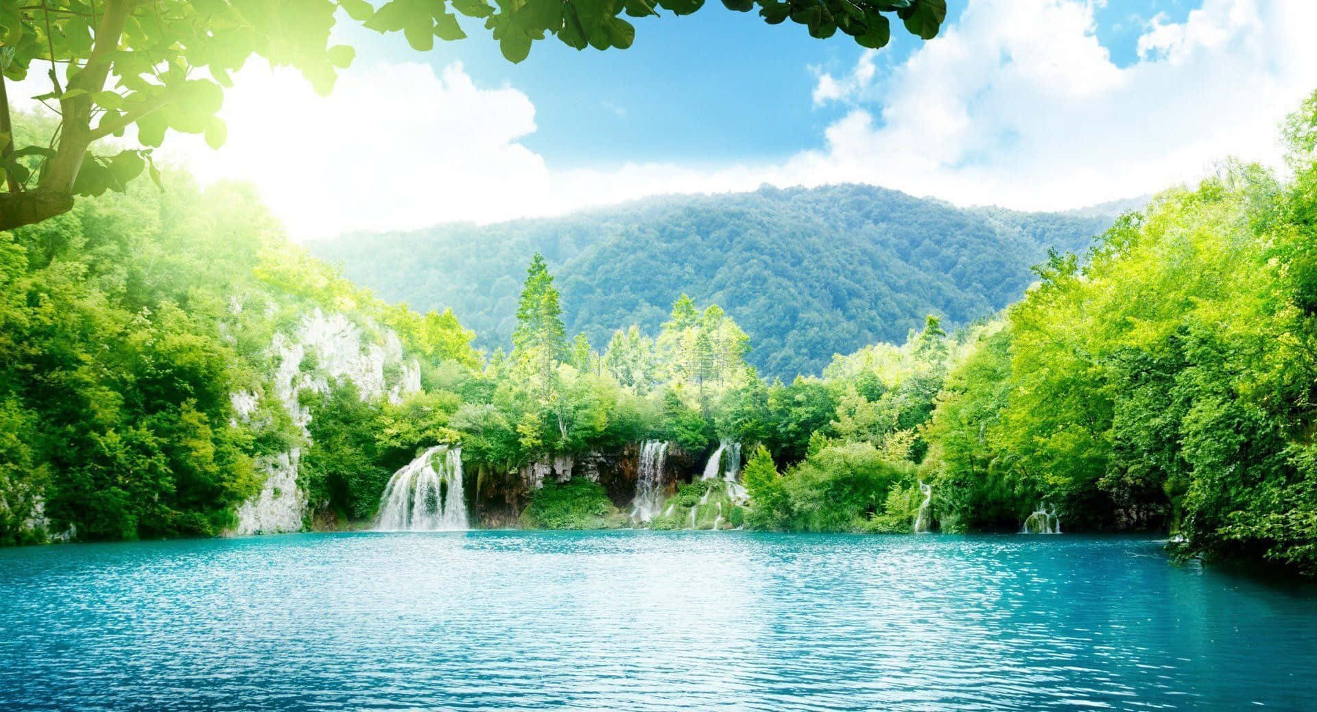 瀑布 桌布 壁紙 — 陽光穿過茂密的綠樹，框住一個寧靜的碧綠湖泊，背景有層疊的瀑布和森林覆蓋的山脈。