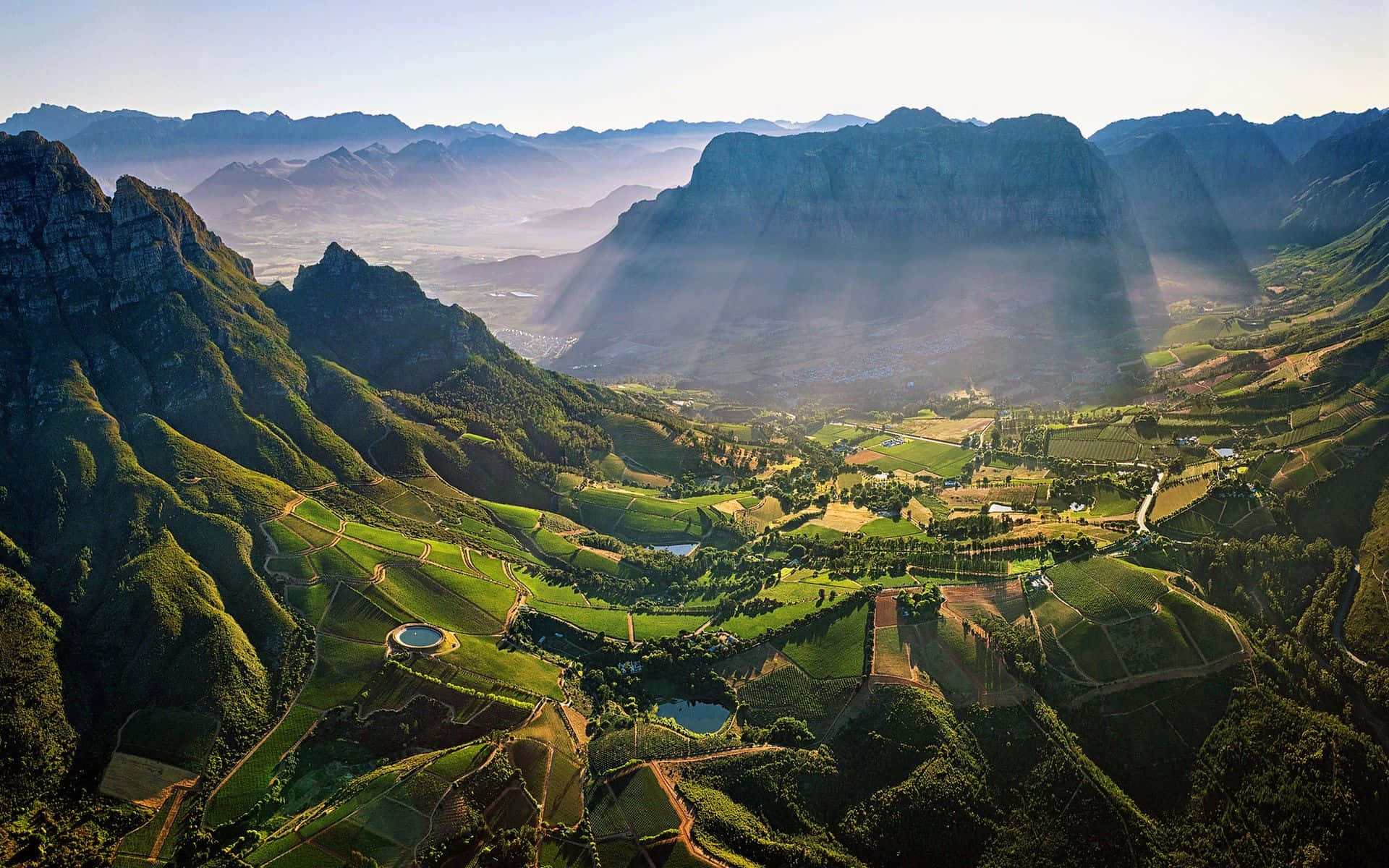 African Landscapes 桌布 壁紙 — 陽光穿過薄霧，照射在層巒疊嶂的山脈之間，一個擁有梯田和小池塘的鬱鬱蔥蔥的綠色山谷。