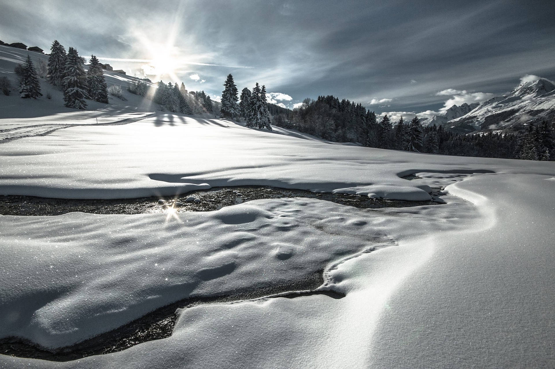 Alps 桌布 壁紙 — 陽光穿過雲層，在白雪覆蓋的山谷上投下長長的陰影，山谷中有蜿蜒的小溪和常綠樹。