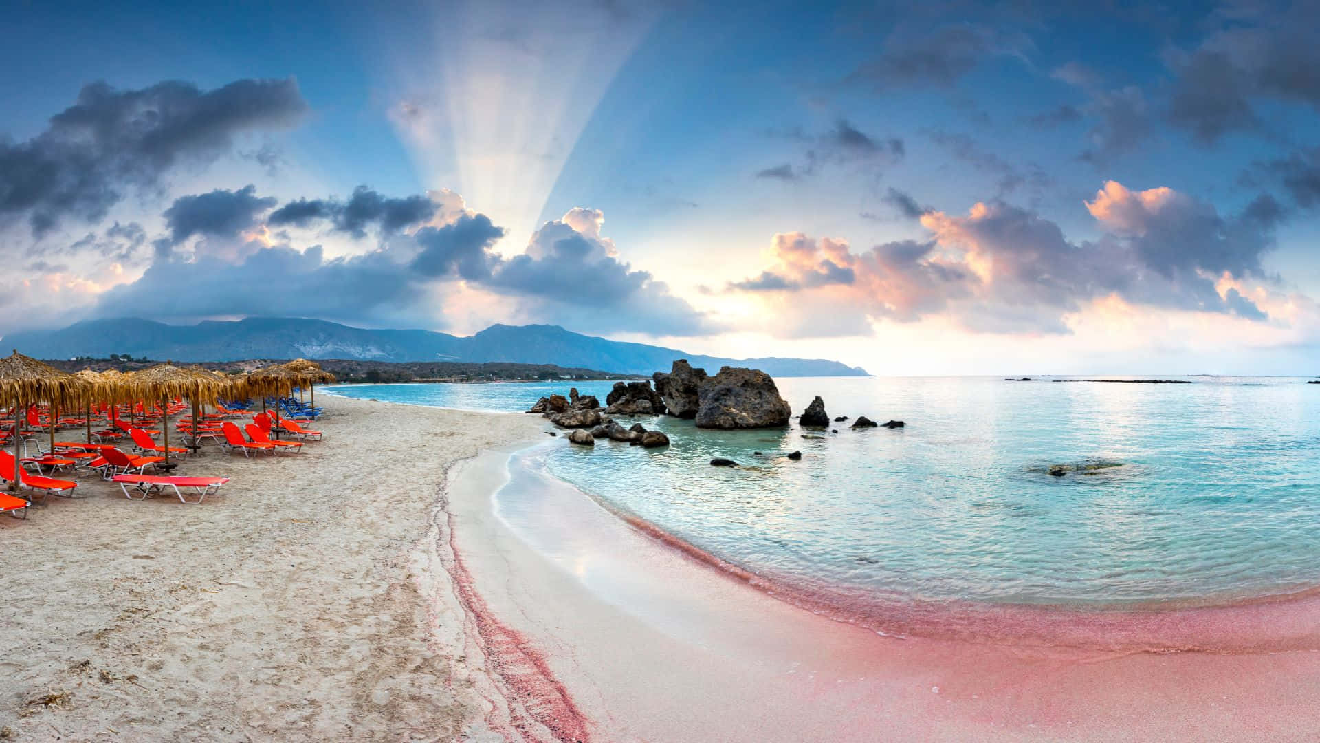 Pink Beach 桌布 壁紙 — 陽光穿過雲層，灑落在一個寧靜的海灘上，沙灘是粉紅色的，海水是碧綠色的，紅色躺椅在草傘下。