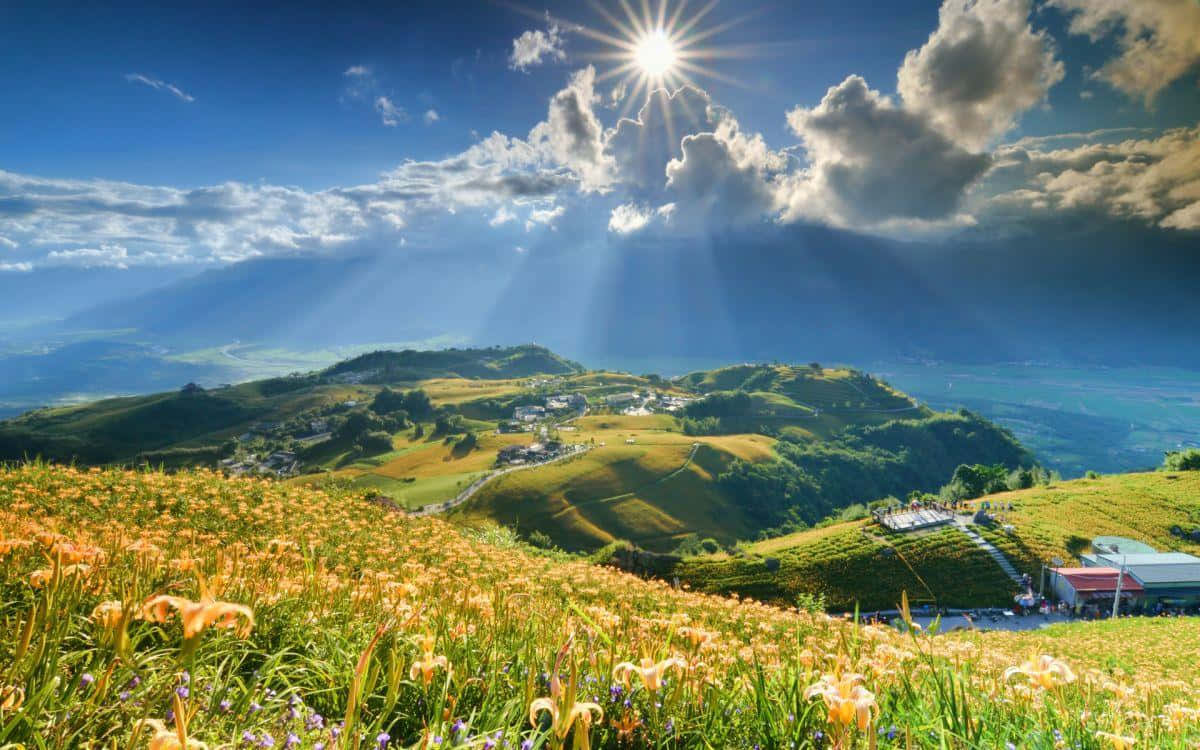 Panoramic Desktop 桌布 壁紙 — 陽光穿過雲層，灑落在起伏的綠色山丘上，山丘上點綴著野花，下方有一個小村莊。