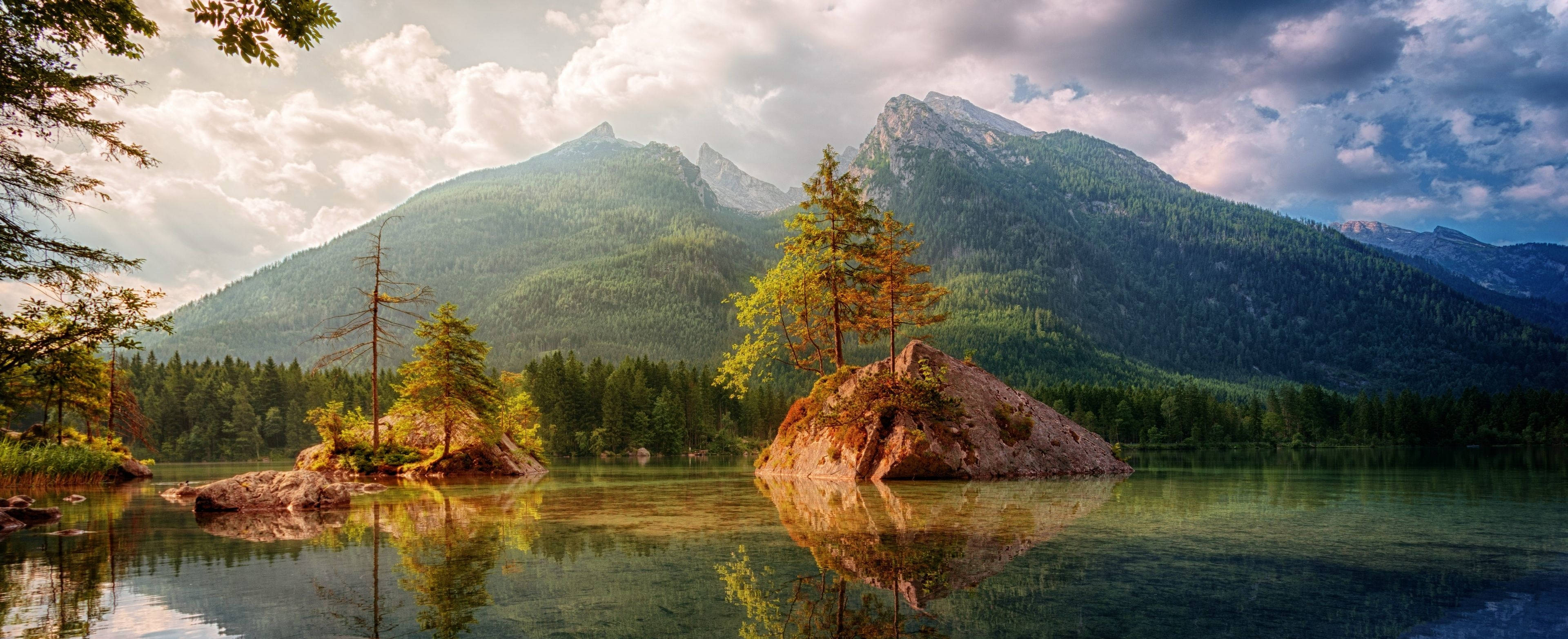 3840x1600 桌布 壁紙 — 陽光穿過雲層，照亮了一個寧靜的山間湖泊，湖中有岩石小島和常綠樹木。