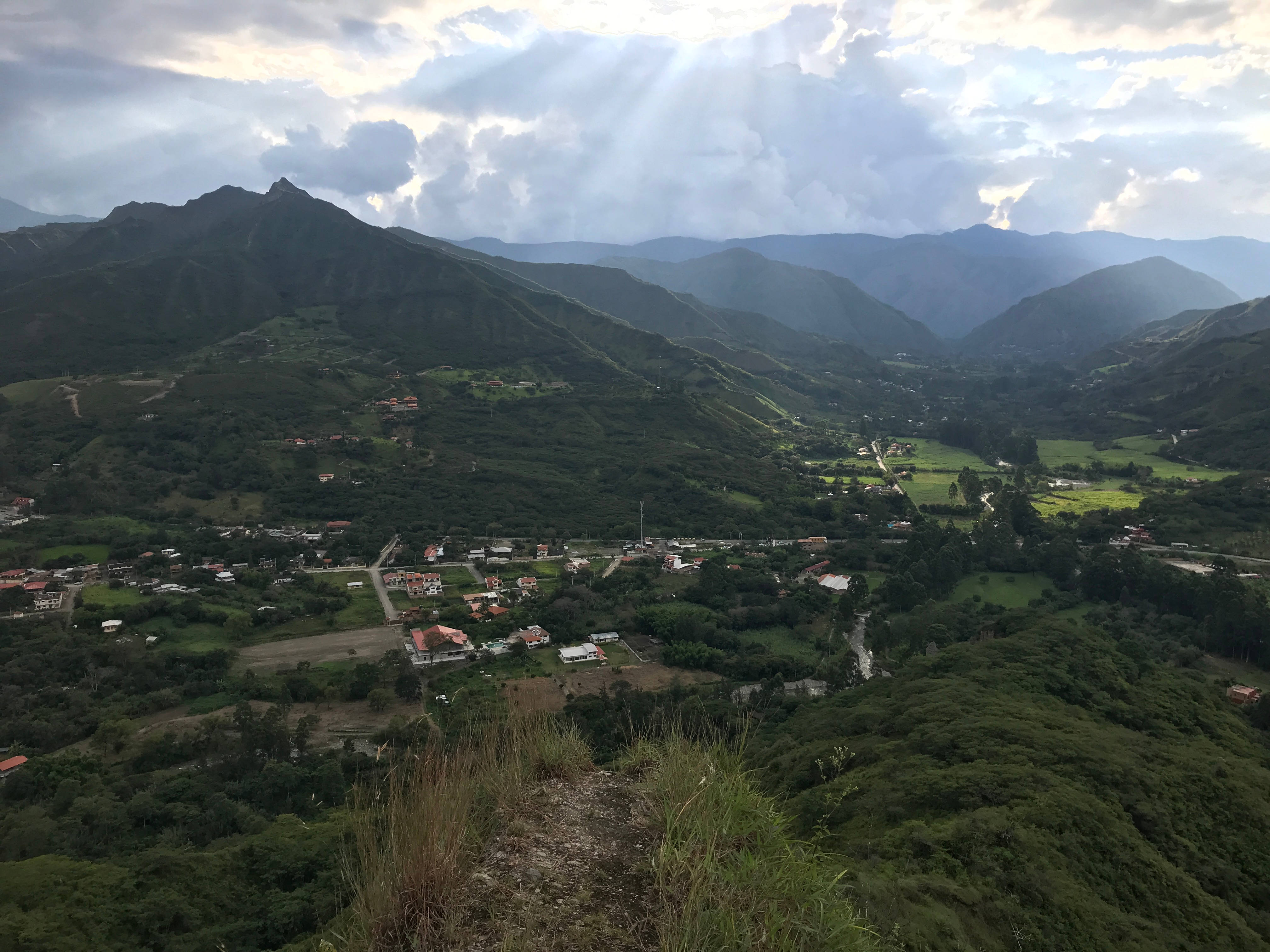 Ecuador 桌布 壁紙 — 陽光穿過雲層，照亮了一個鬱鬱蔥蔥的綠色山谷，山谷中有一個小村莊坐落在連綿起伏的山巒之間。