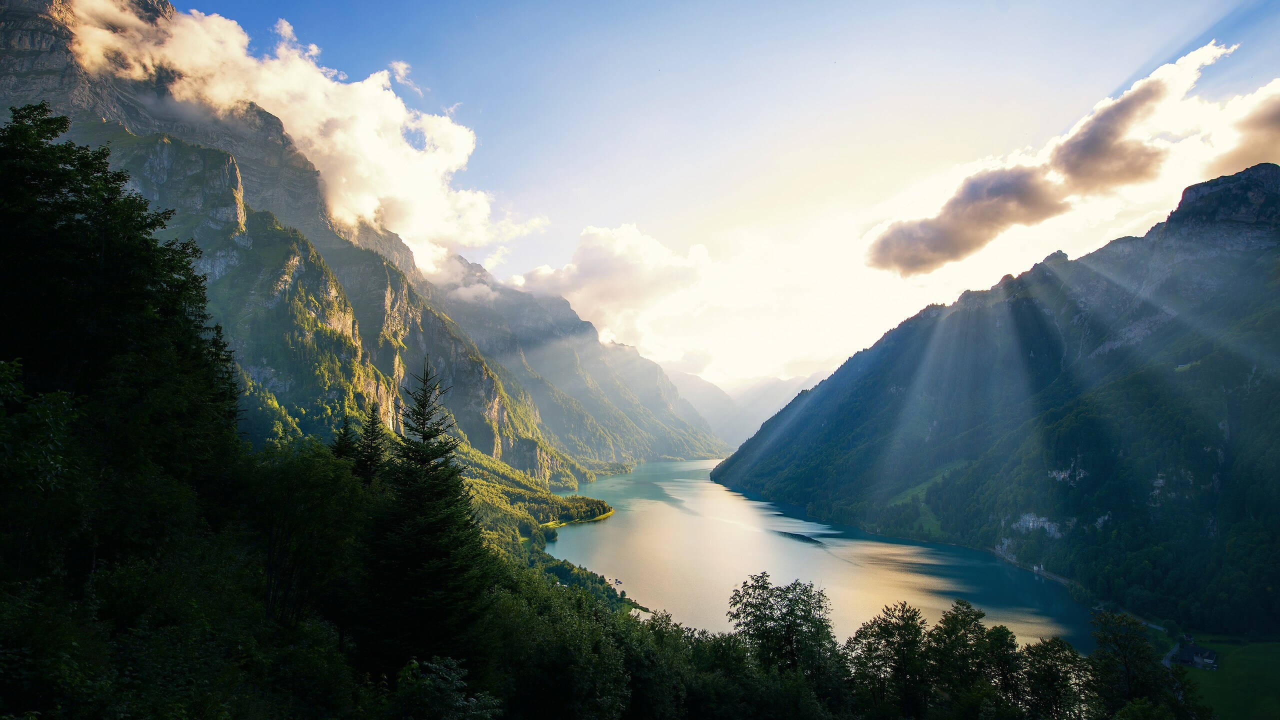 2560x1440 nature 桌布 壁紙 — 陽光穿過雲層，照亮了坐落在高聳翠綠山巒之間的寧靜高山湖泊。