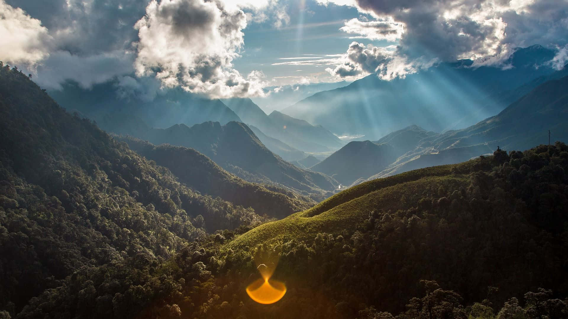 風景 桌布 壁紙 — 陽光穿過雲層，照亮了覆蓋著茂密綠色森林的層層山脈。