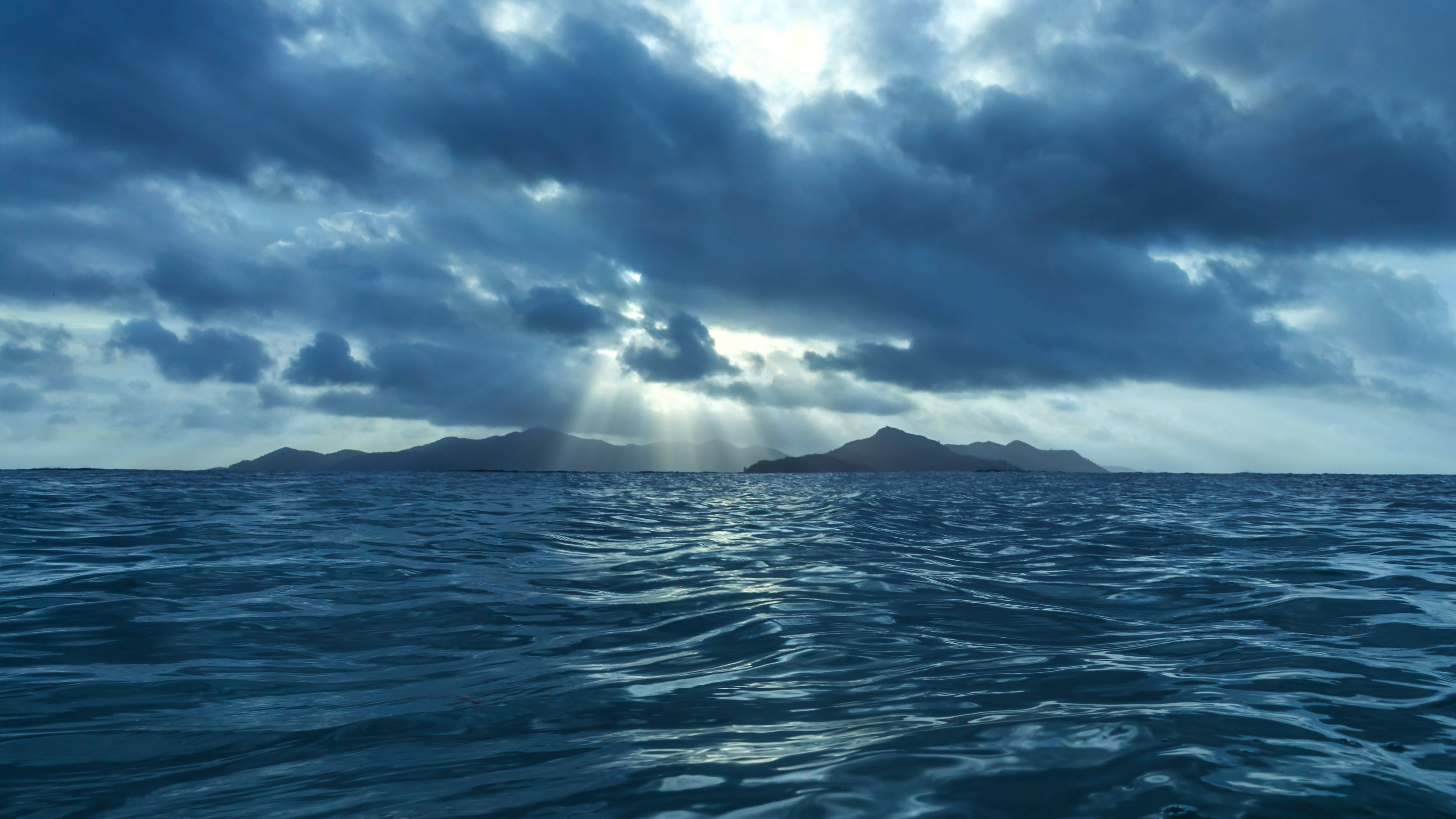 海洋 桌布 壁紙 — 陽光穿過黑暗的暴風雲，照耀在廣闊的海洋上，遠方地平線上有著多山的島嶼。