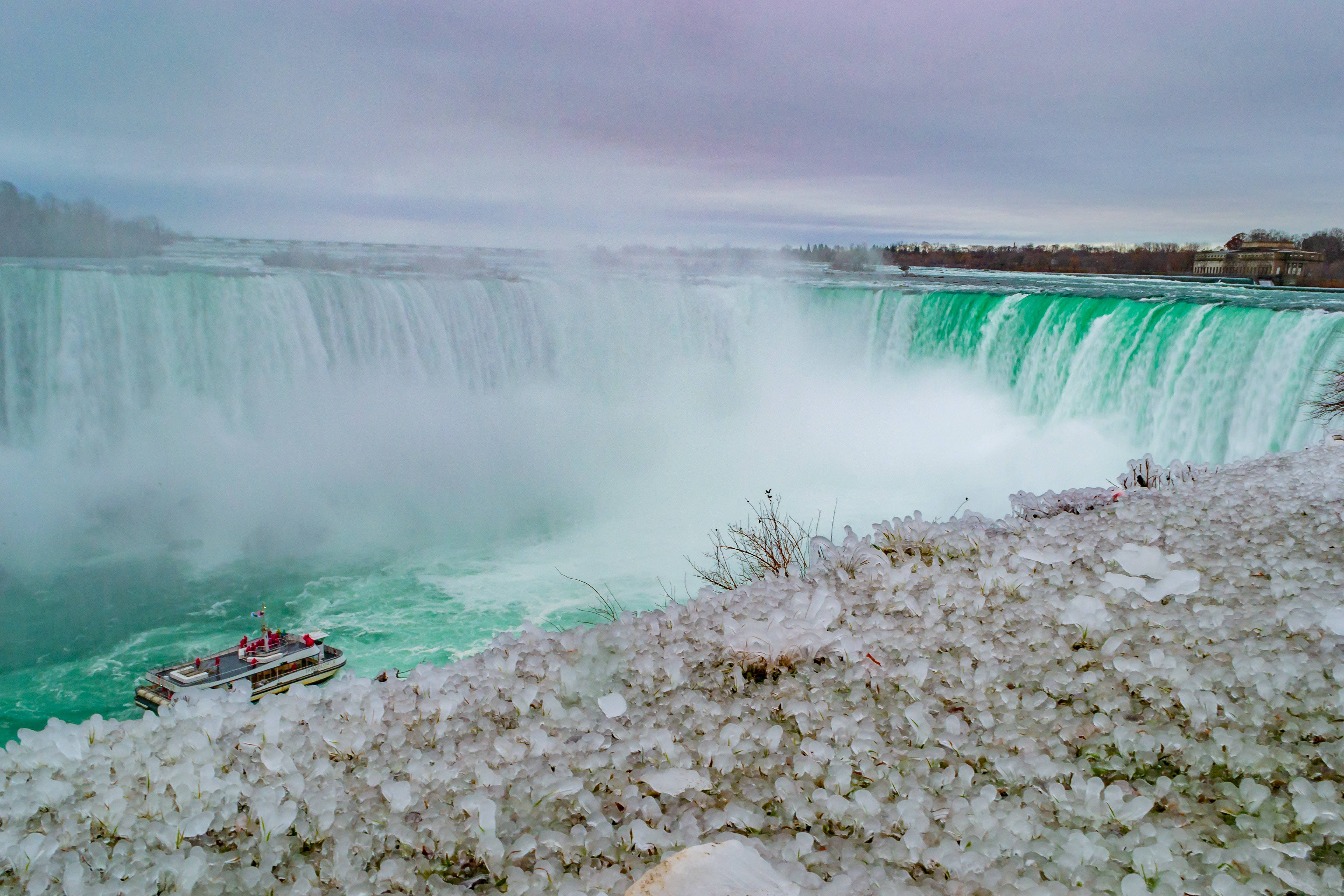 niagara 桌布 壁紙 — 雄偉的冬季瀑布傾瀉在碧綠色的水中，前景有冰晶排列，一艘觀光船在霧氣瀰漫的水域中航行。