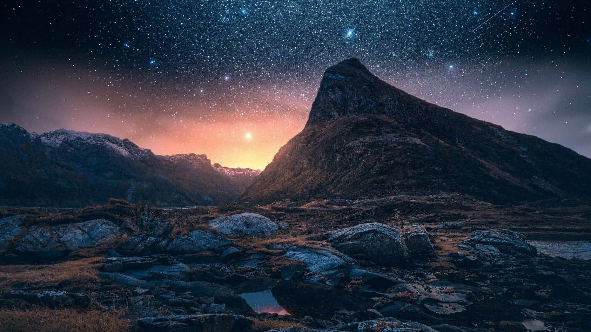 山景 桌布 壁紙 — 雄偉的山峰在繁星點點的夜空下升起，地平線上閃耀的日出映照在岩石前景上。