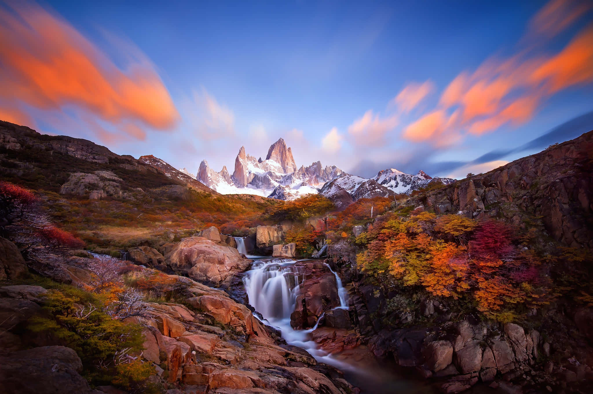 Patagonia 桌布 壁紙 — 雄偉的山脈，山頂積雪，在層疊瀑布後方升起，周圍環繞著秋葉，天空呈現鮮豔的橙色和藍色。