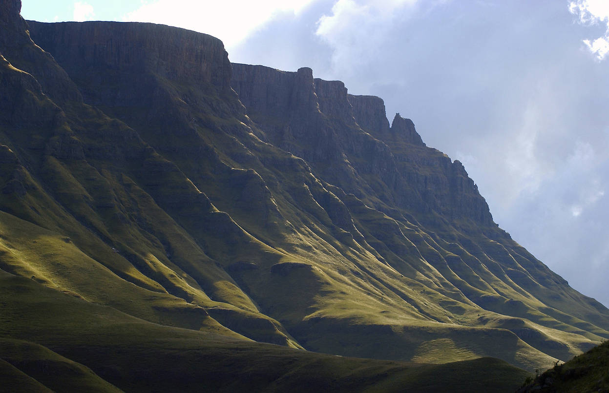 Lesotho 桌布 壁紙 — 雄偉的山脈，陡峭的草坡沐浴在金色的陽光下，映襯著多雲的天空。