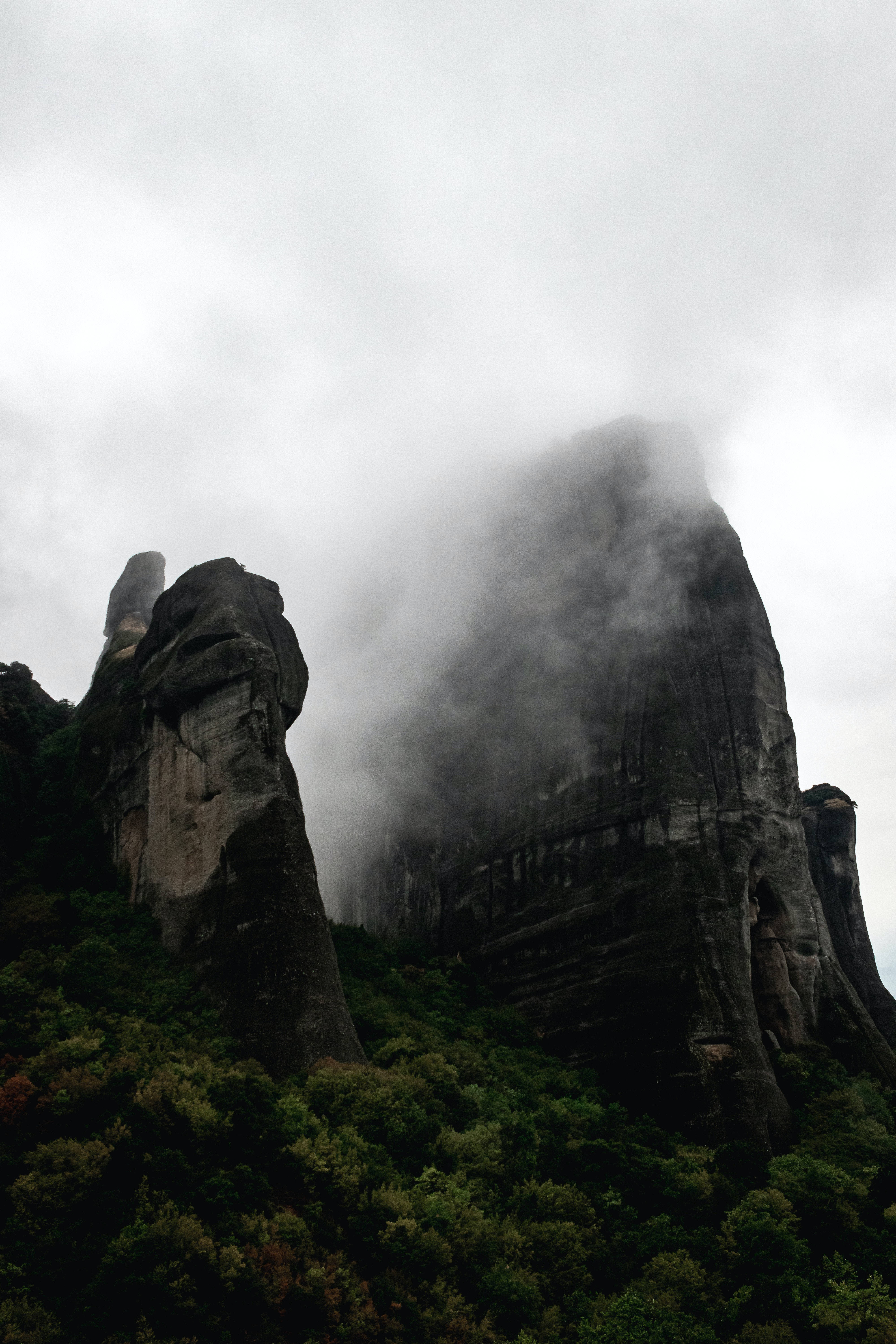雲朵 桌布 壁紙 — 雄偉的深色岩石從茂密的綠色森林中升起，霧氣在陰天籠罩下繞著山頂盤旋。