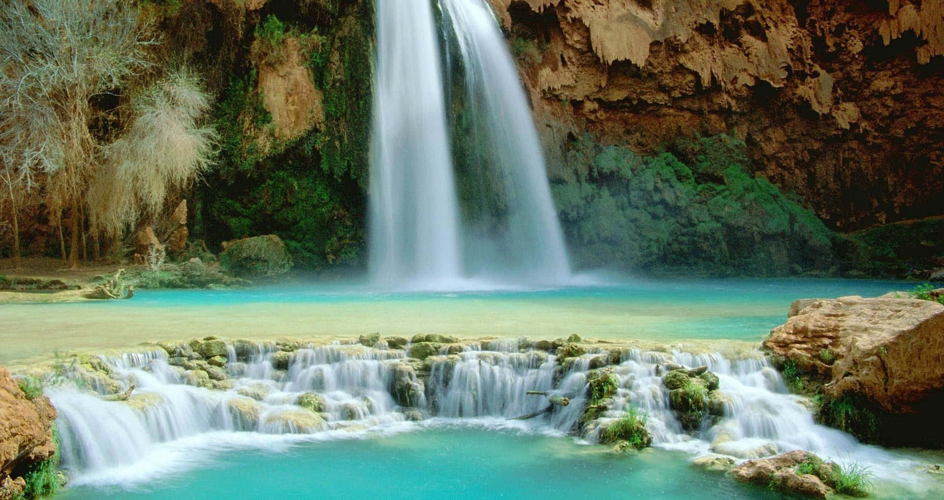 Waterfalls 桌布 壁紙 — 雄偉的瀑布傾瀉而下，流入充滿活力的綠松石色水池，四周環繞著茂密的植被和崎嶇的懸崖。