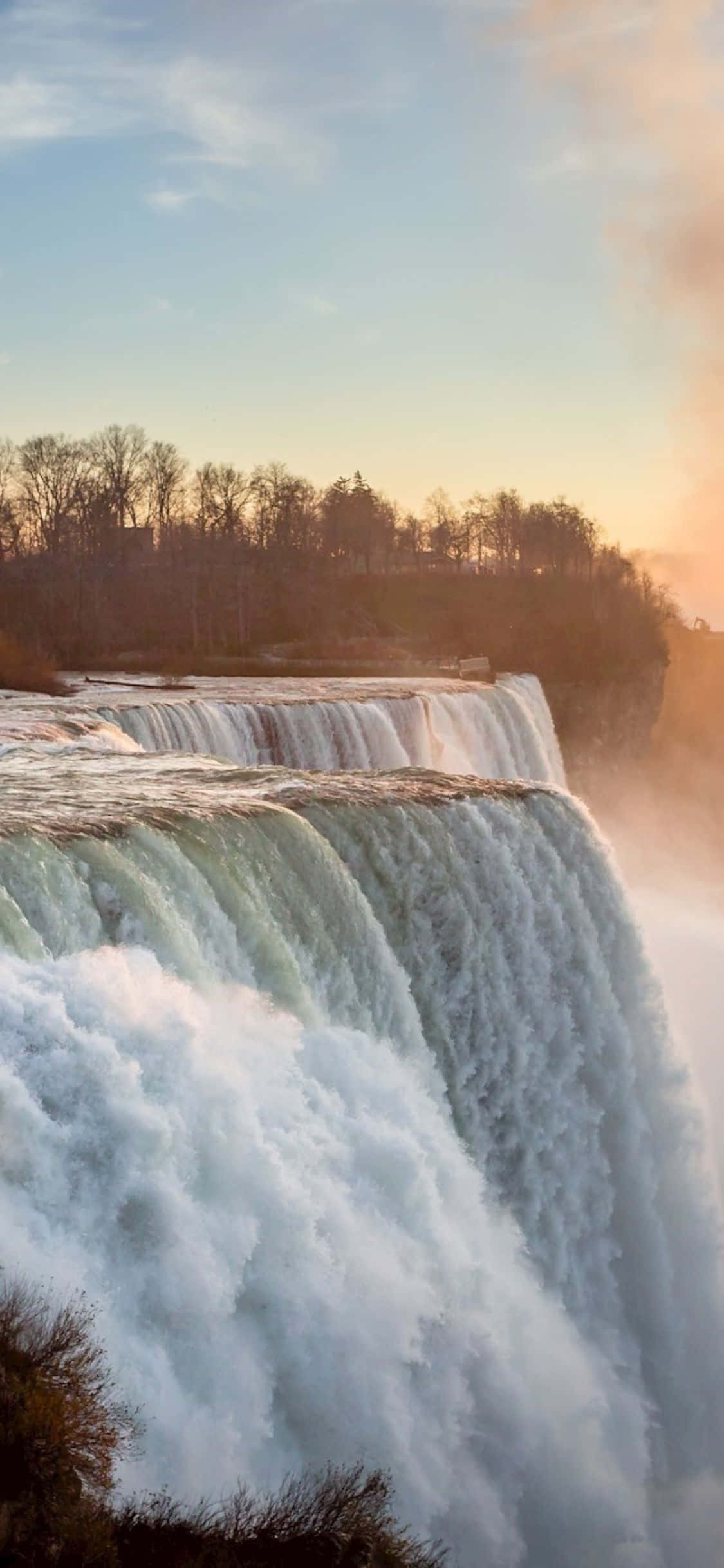 Niagara Falls 桌布 壁紙 — 雄偉的瀑布在日落時分強力傾瀉而下，霧氣升騰，光禿的樹木在溫暖的天空襯托下形成剪影。