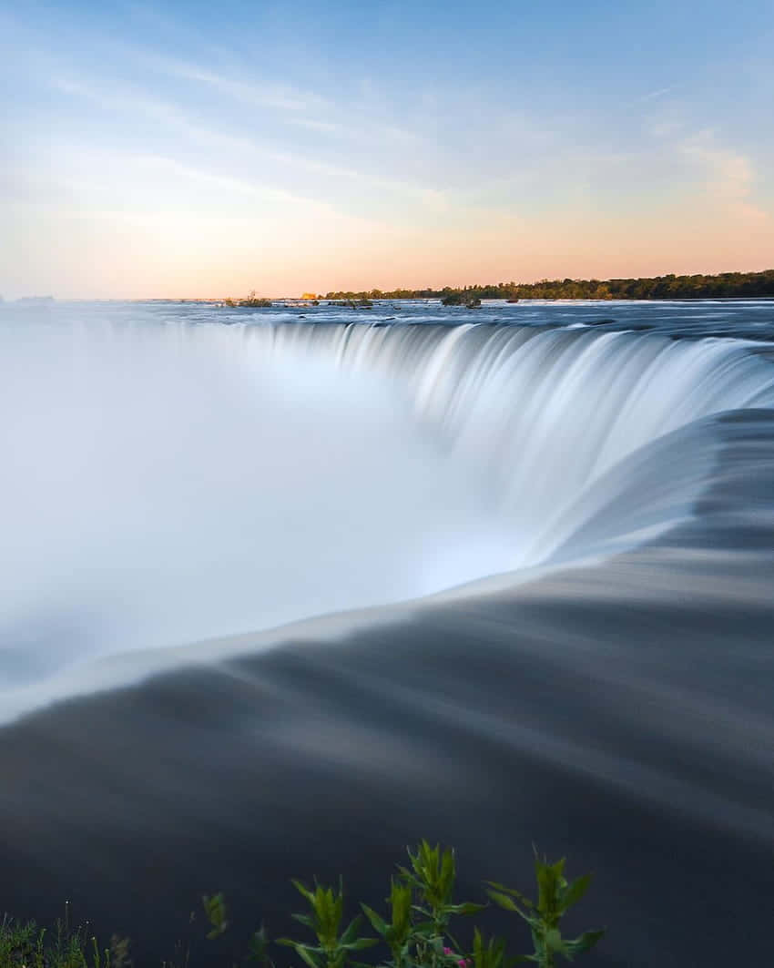 720p Niagara Falls 桌布 壁紙 — 雄偉的瀑布在柔和的日出天空下以慢動作傾瀉而下，薄霧從深色水面上昇起，前景有綠色植物。