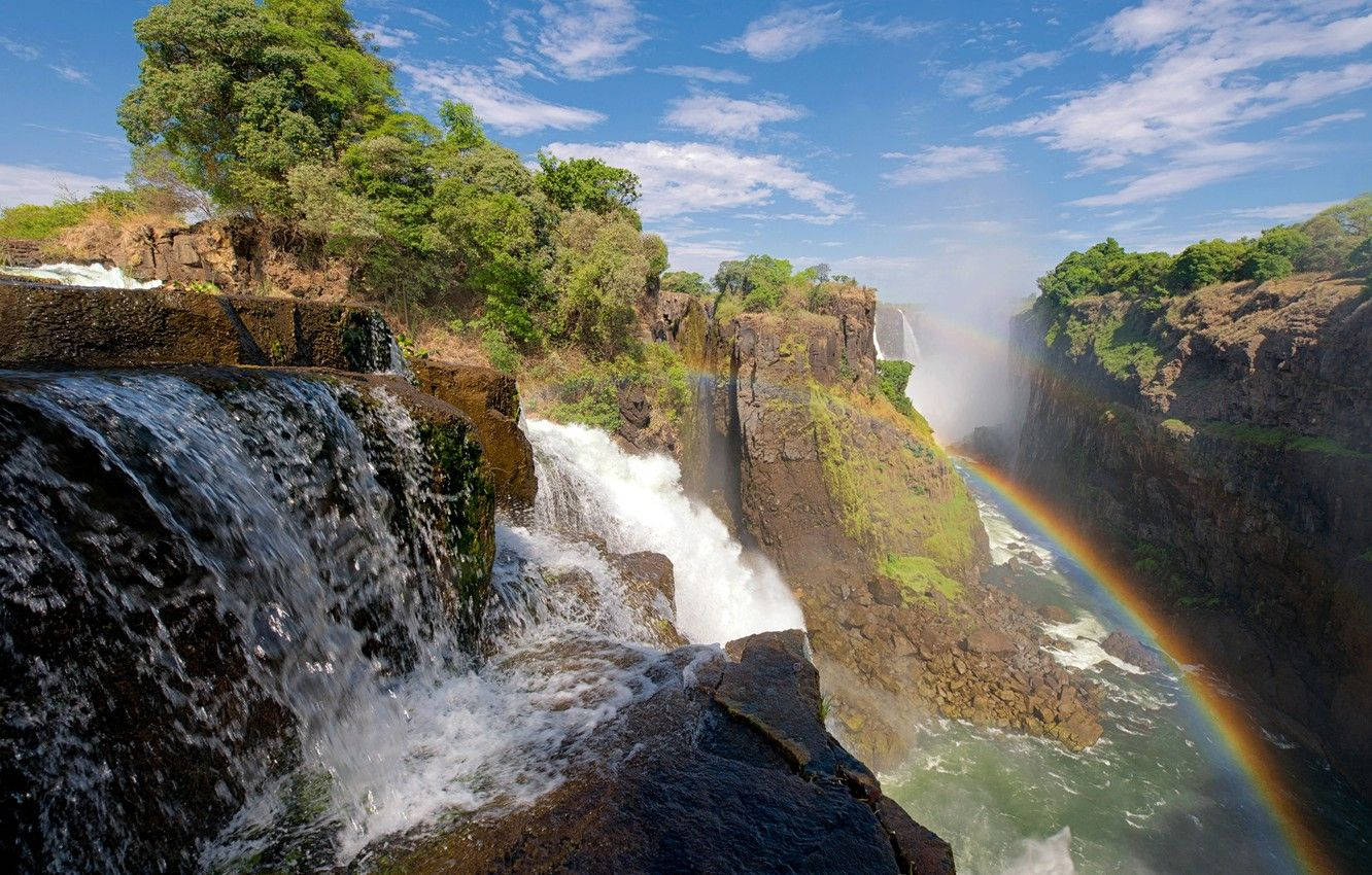 Zimbabwe 桌布 壁紙 — 雄偉的瀑布從岩石懸崖上傾瀉而下，在晴朗的藍天和白雲下形成一道鮮豔的彩虹。
