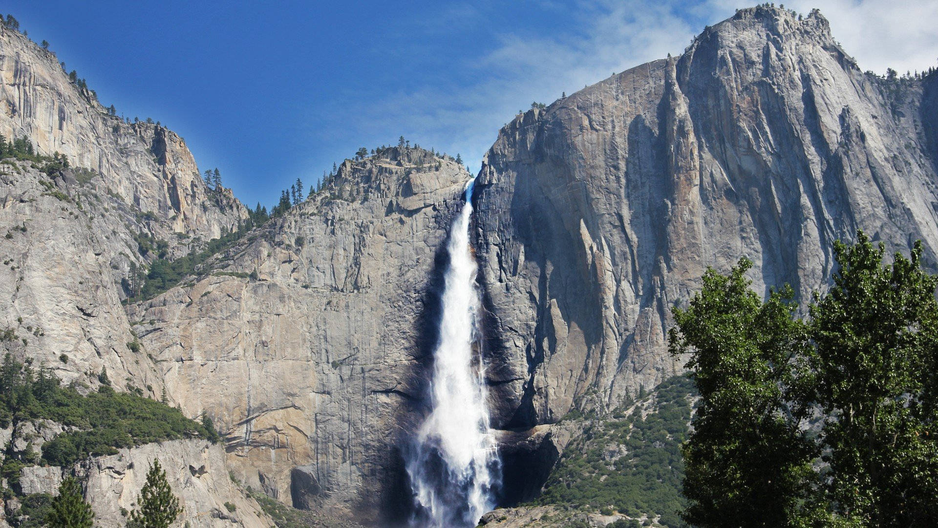 Falling Water 桌布 壁紙 — 雄偉的瀑布從高聳的花崗岩懸崖傾瀉而下，在清澈的藍天下，茂密的綠樹環繞著整個場景。