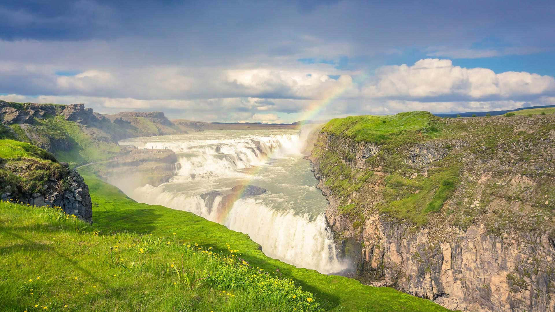 Iceland 桌布 壁紙 — 雄偉的瀑布穿過深邃的峽谷，在部分多雲的天空下，一道鮮豔的彩虹拱在鬱鬱蔥蔥的綠色懸崖上。