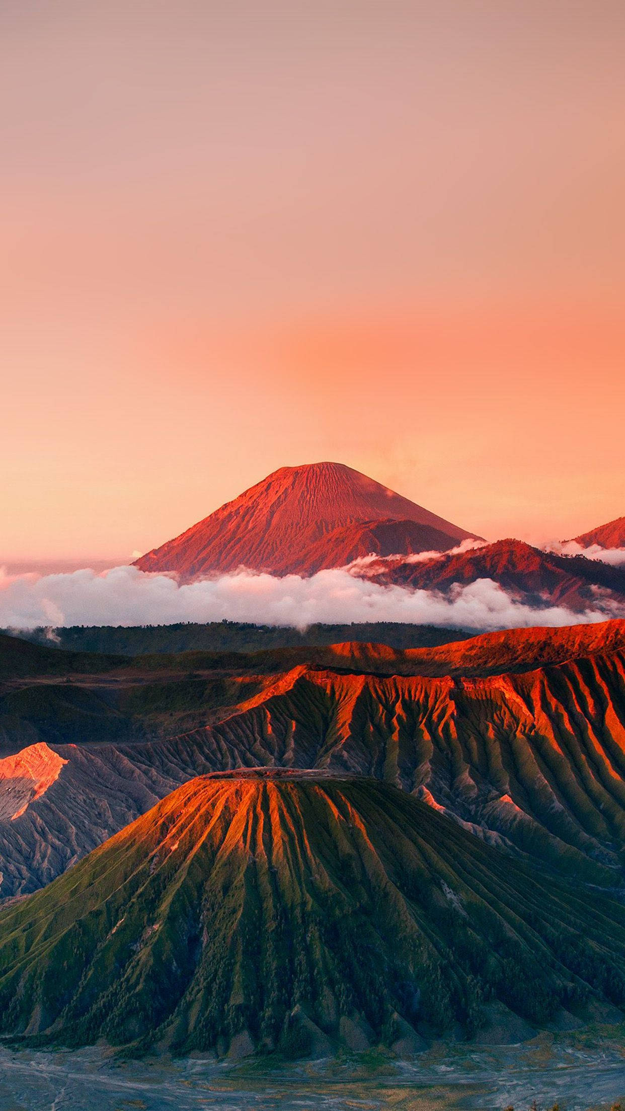 紅色山脈 桌布 壁紙 — 雄偉的火山山峰在日出時聳立於雲海之上，溫暖的橙色和粉紅色調沐浴著崎嶇的地形。